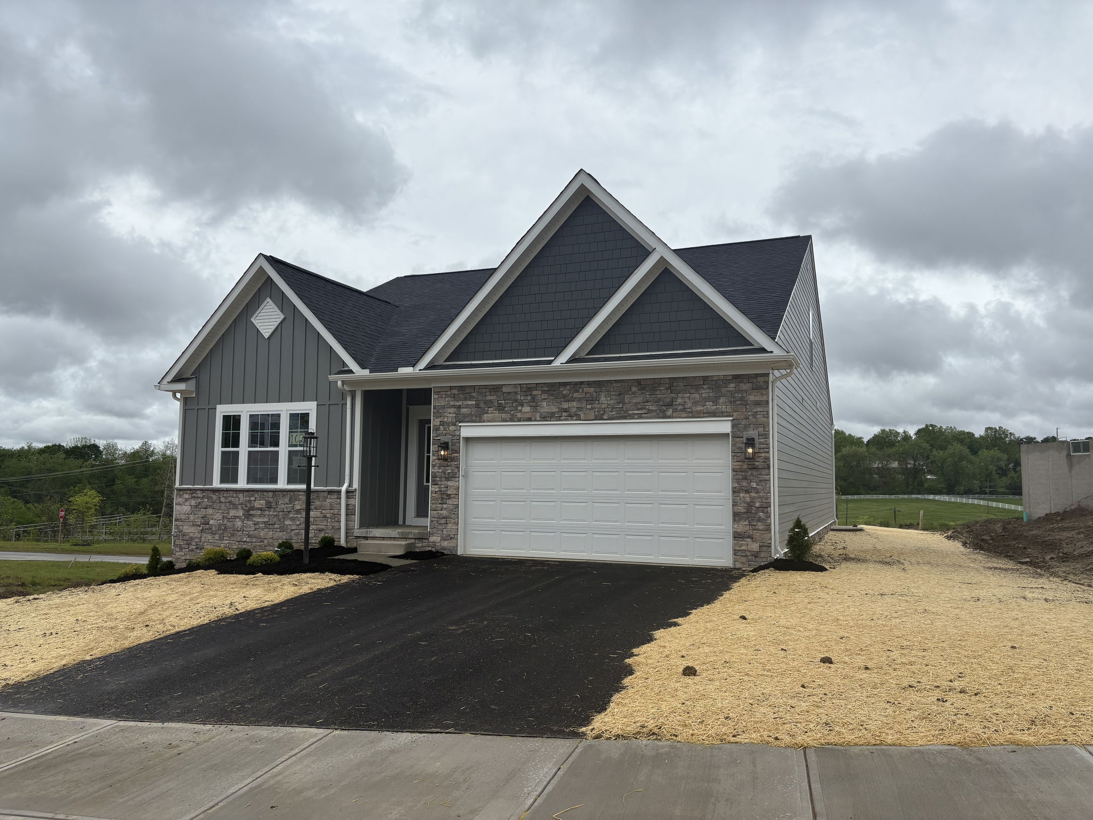 Modern gray brick house with a dark shingled roof, two-car garage, newly paved driveway, and freshly seeded lawn under a cloudy sky.