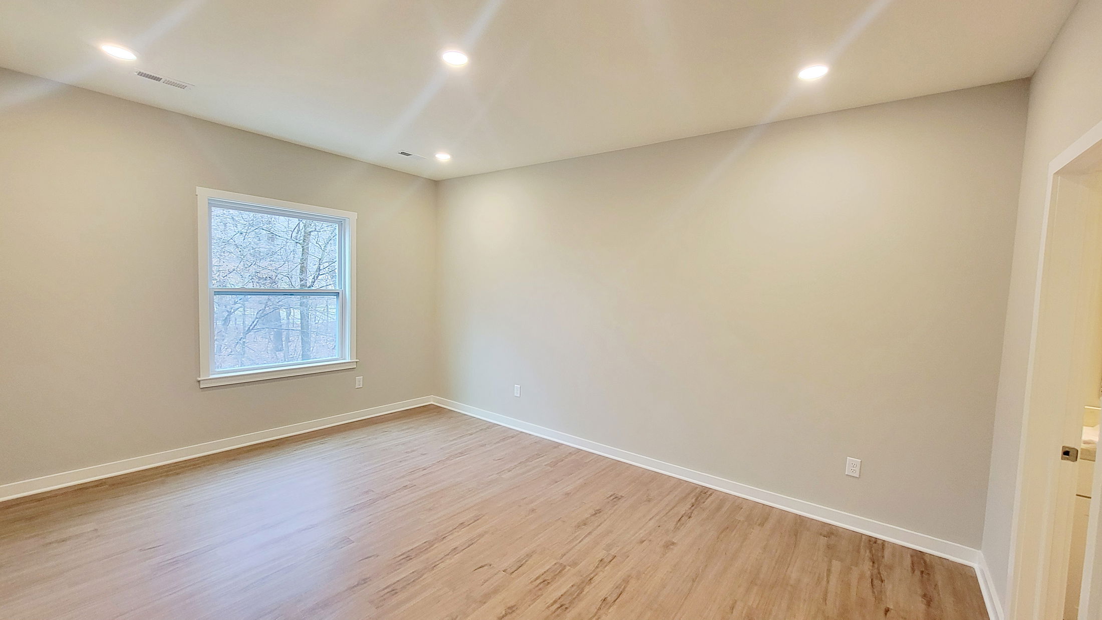 Spacious empty room with light wooden flooring, beige walls, and a large window.
