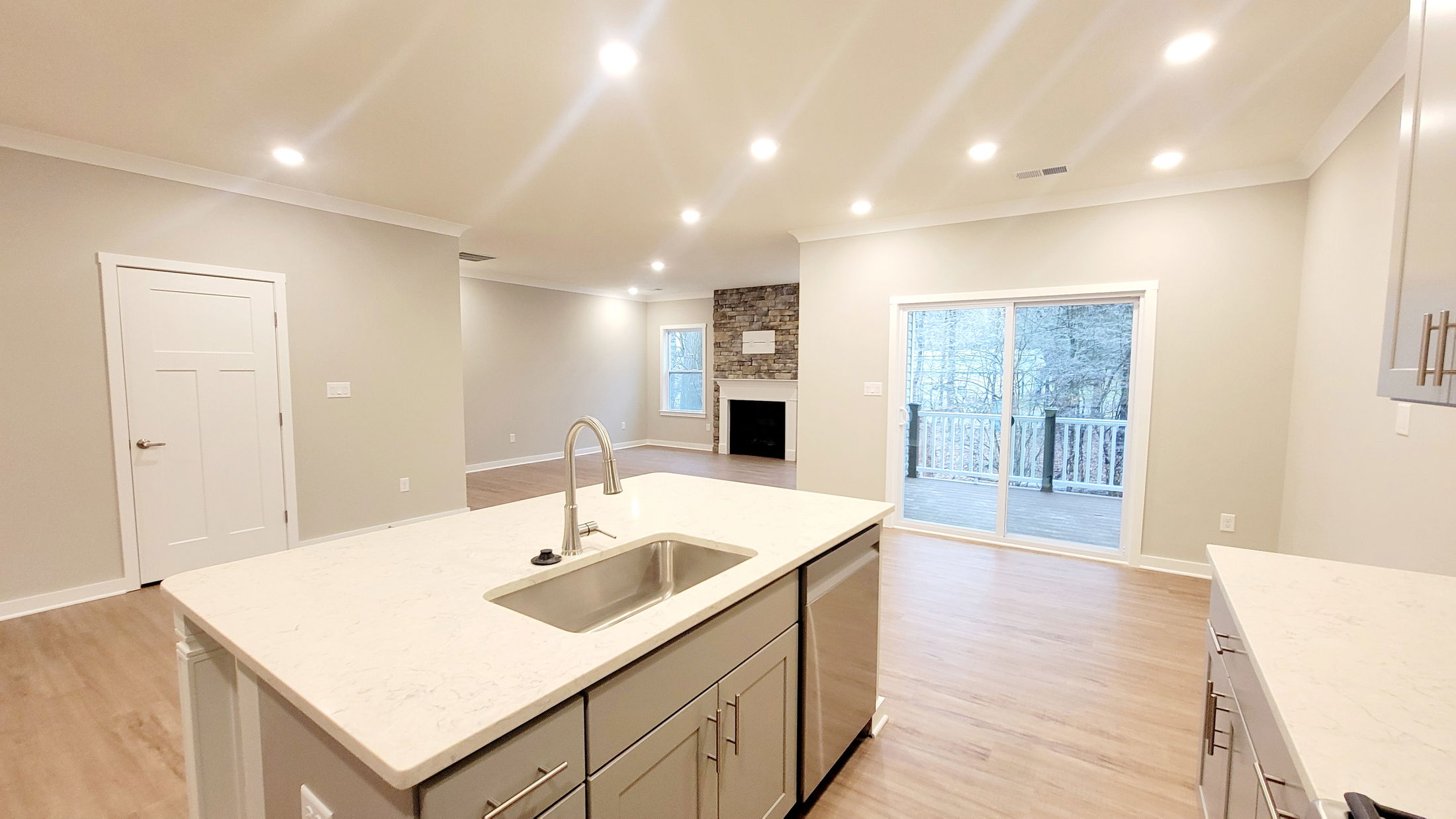 Bright and spacious modern kitchen and living room with an island, hardwood floors, stone fireplace, and sliding glass doors leading to a deck.