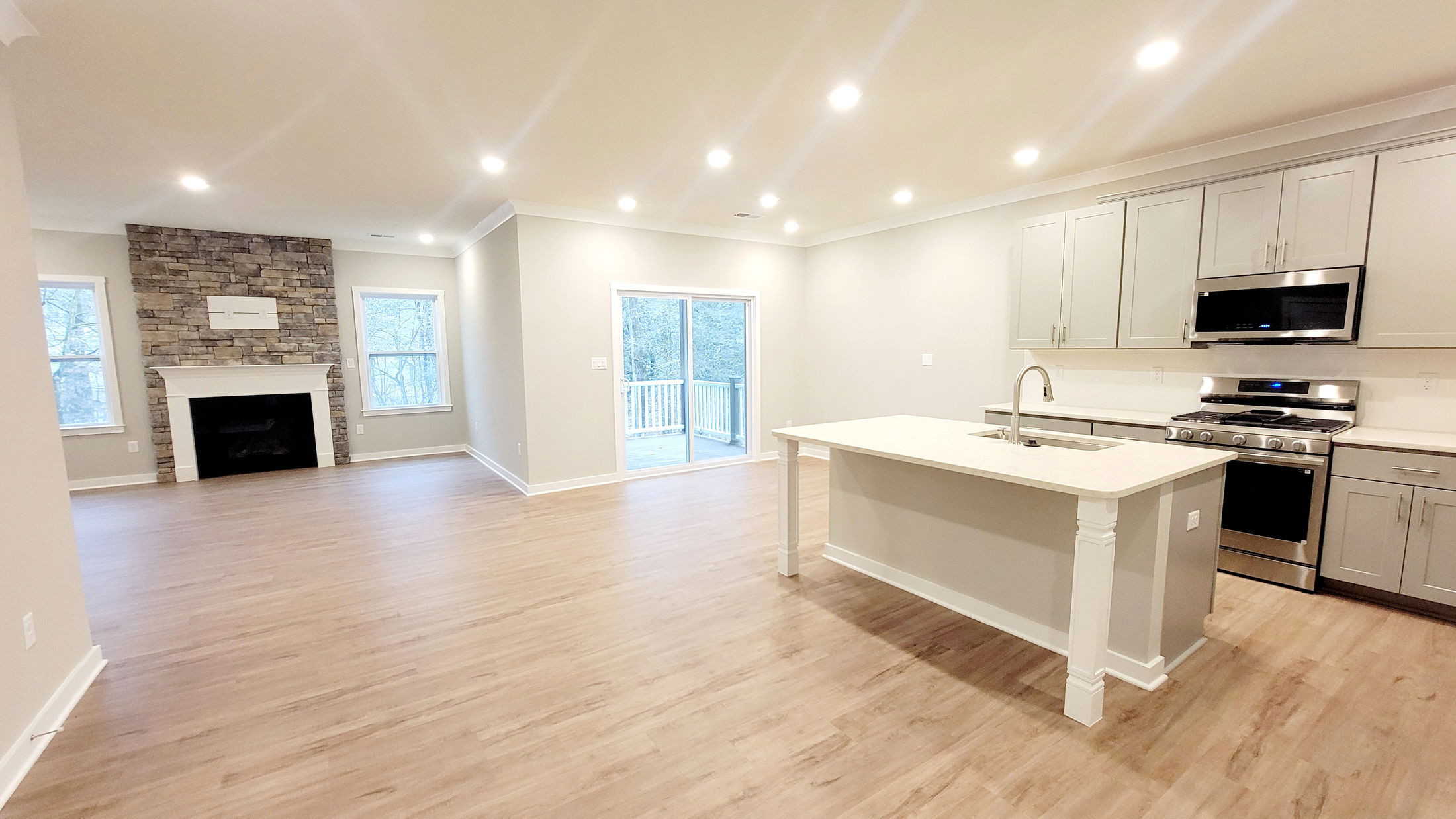Modern open-concept kitchen and living area with hardwood floors, stone fireplace, and large glass doors leading to a deck.