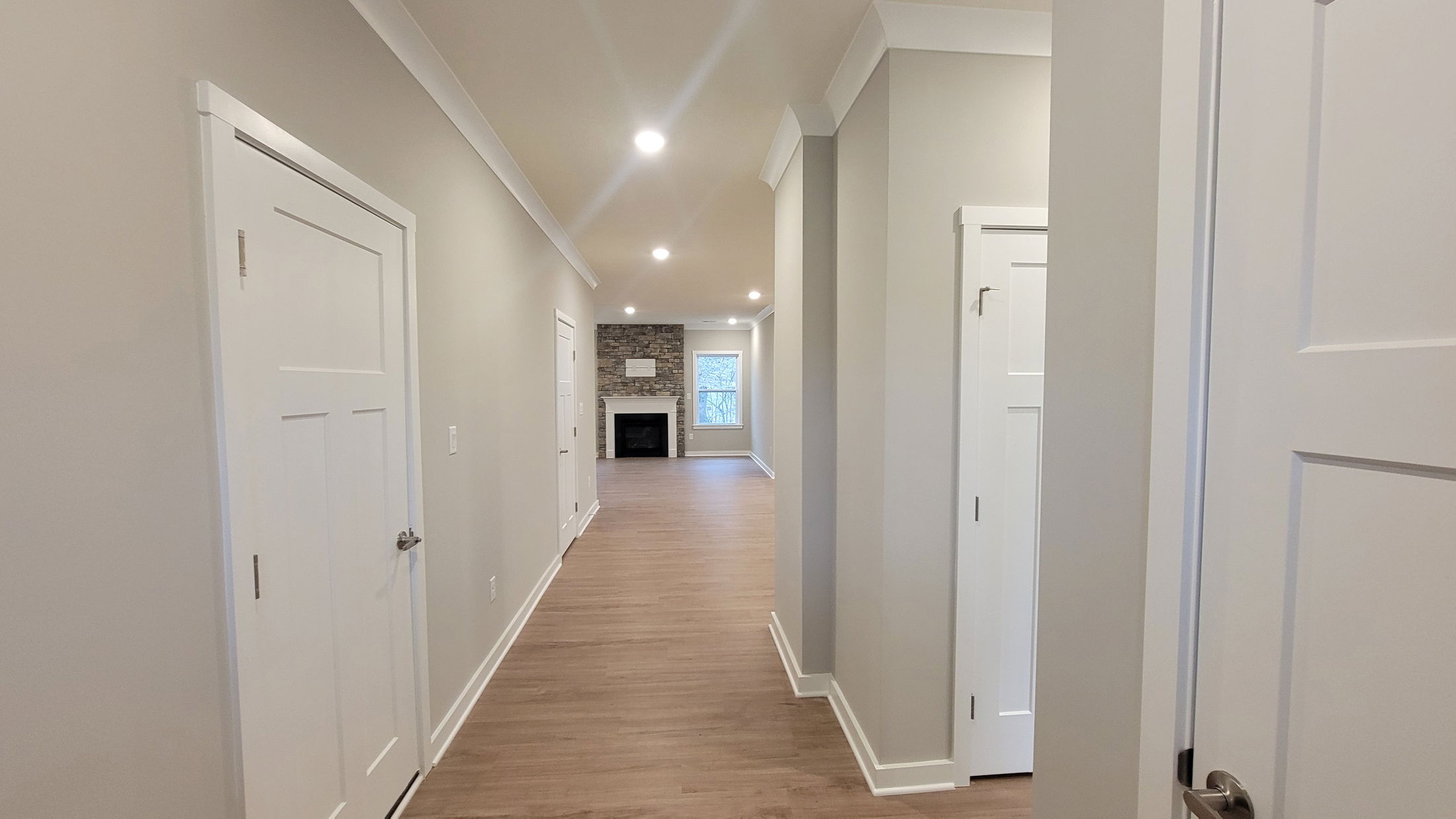 Modern hallway with light wood flooring, leading to a stone fireplace and large window in a spacious living area.