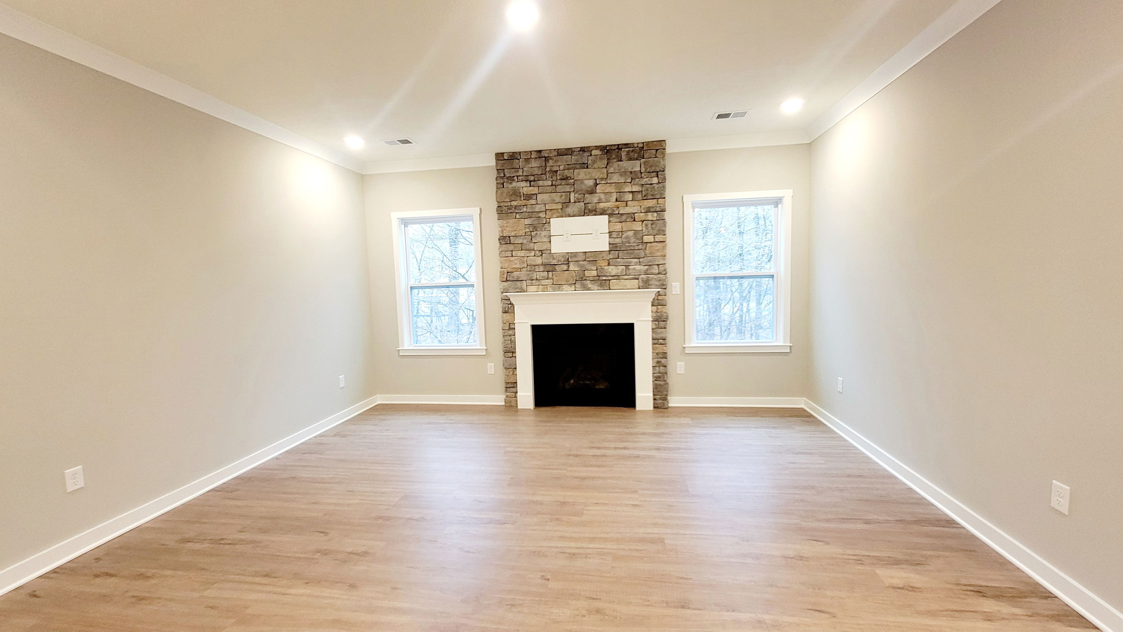 Spacious living room with light wood flooring, neutral walls, and a stone fireplace flanked by two large windows.