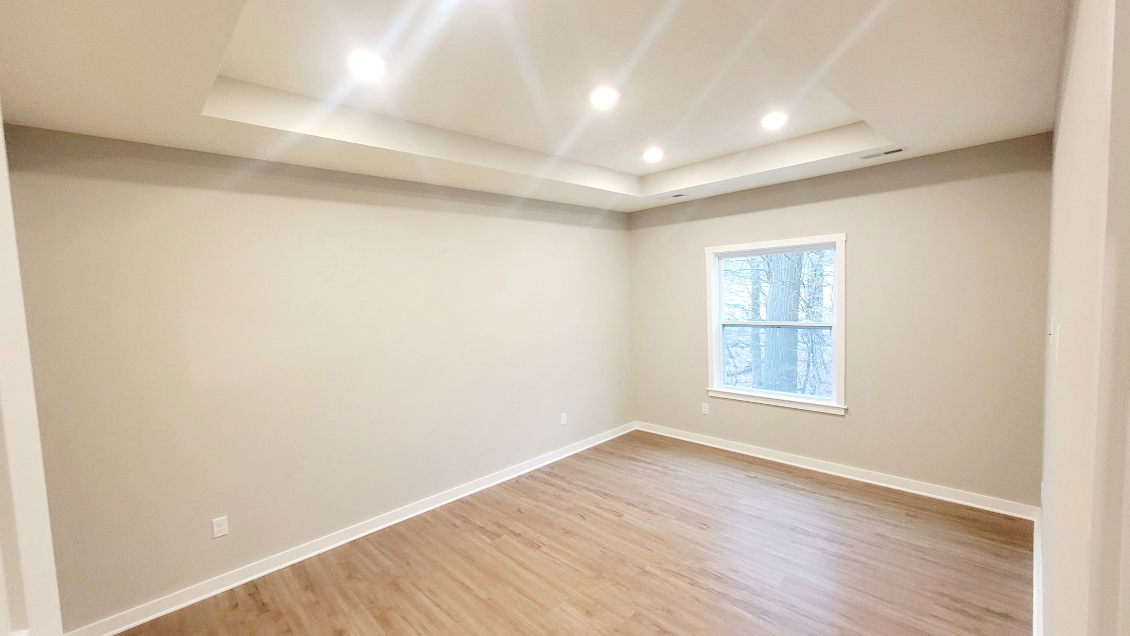 Bright, empty room with recessed lighting, beige walls, and a large window overlooking trees.