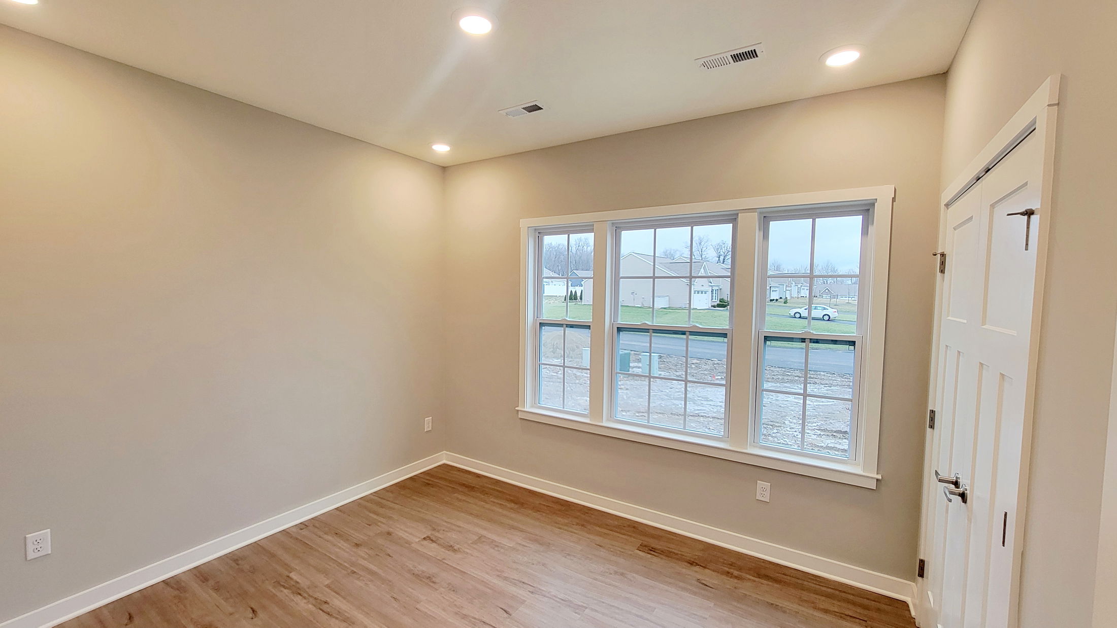 Empty bedroom with light wood flooring, beige walls, large windows, and recessed ceiling lights.