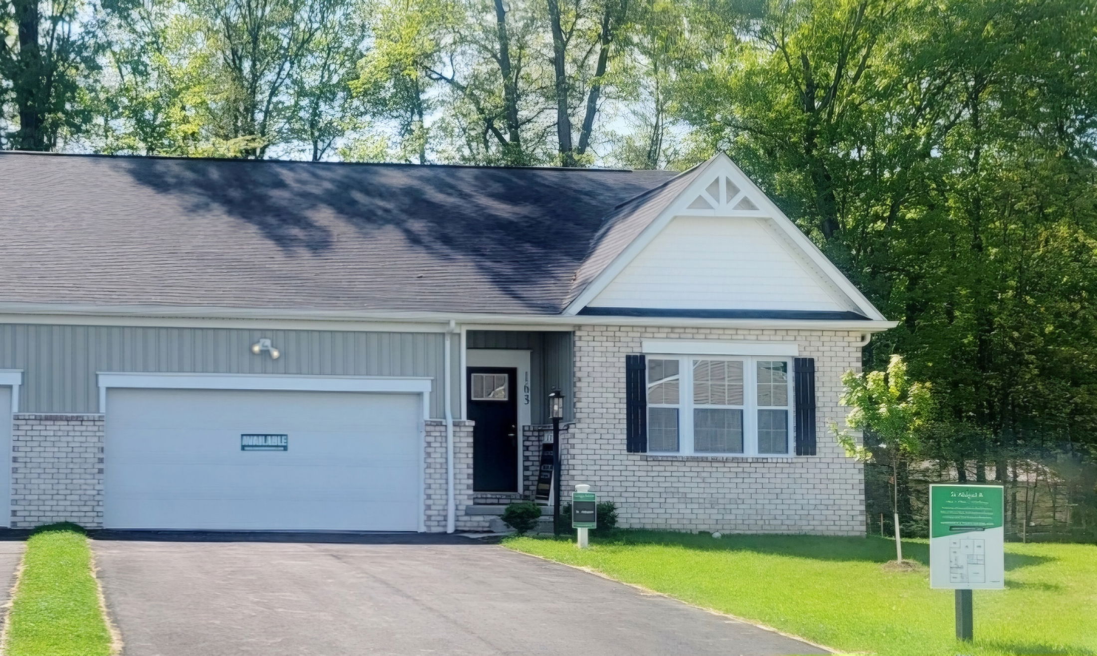Single-story brick house with a two-car garage, dark shutters, and lush green lawn, nestled by tall trees.