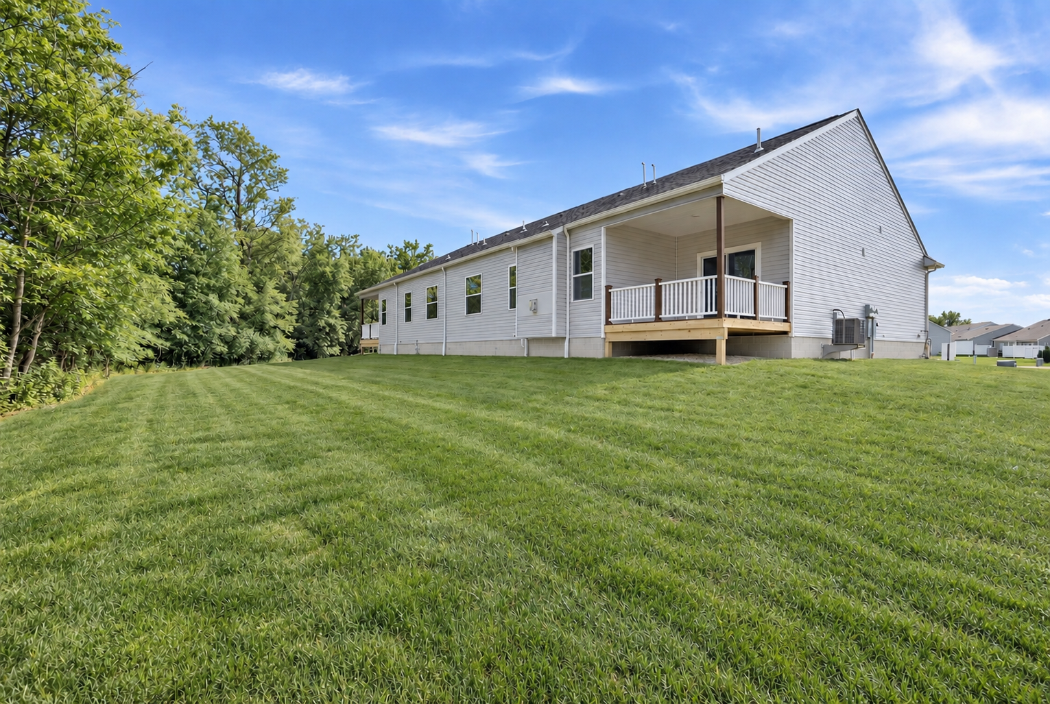 A white house with a wooden deck sits on a large, well-maintained grassy yard surrounded by trees under a blue sky.