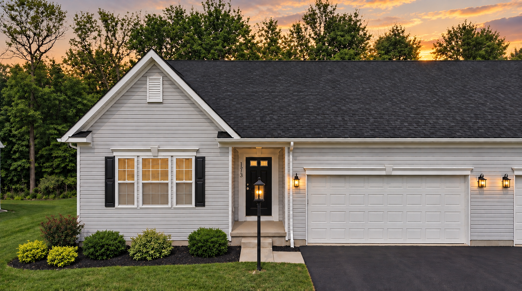 Single-story gray house with a black roof, white trim, landscaped yard, and a double garage under a sunset sky.