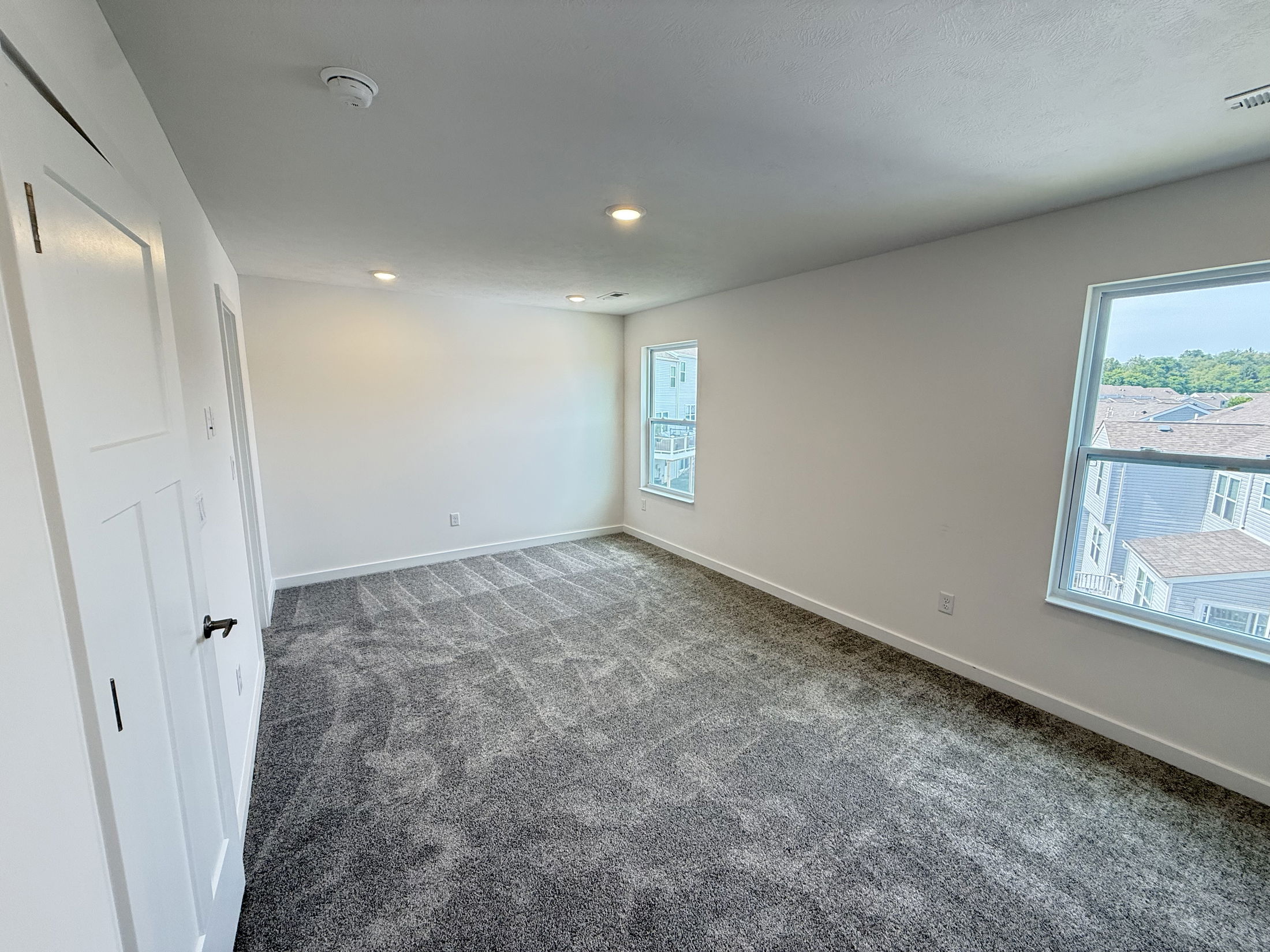 Spacious, empty interior room with gray carpet, bright white walls, and large windows letting in natural light.