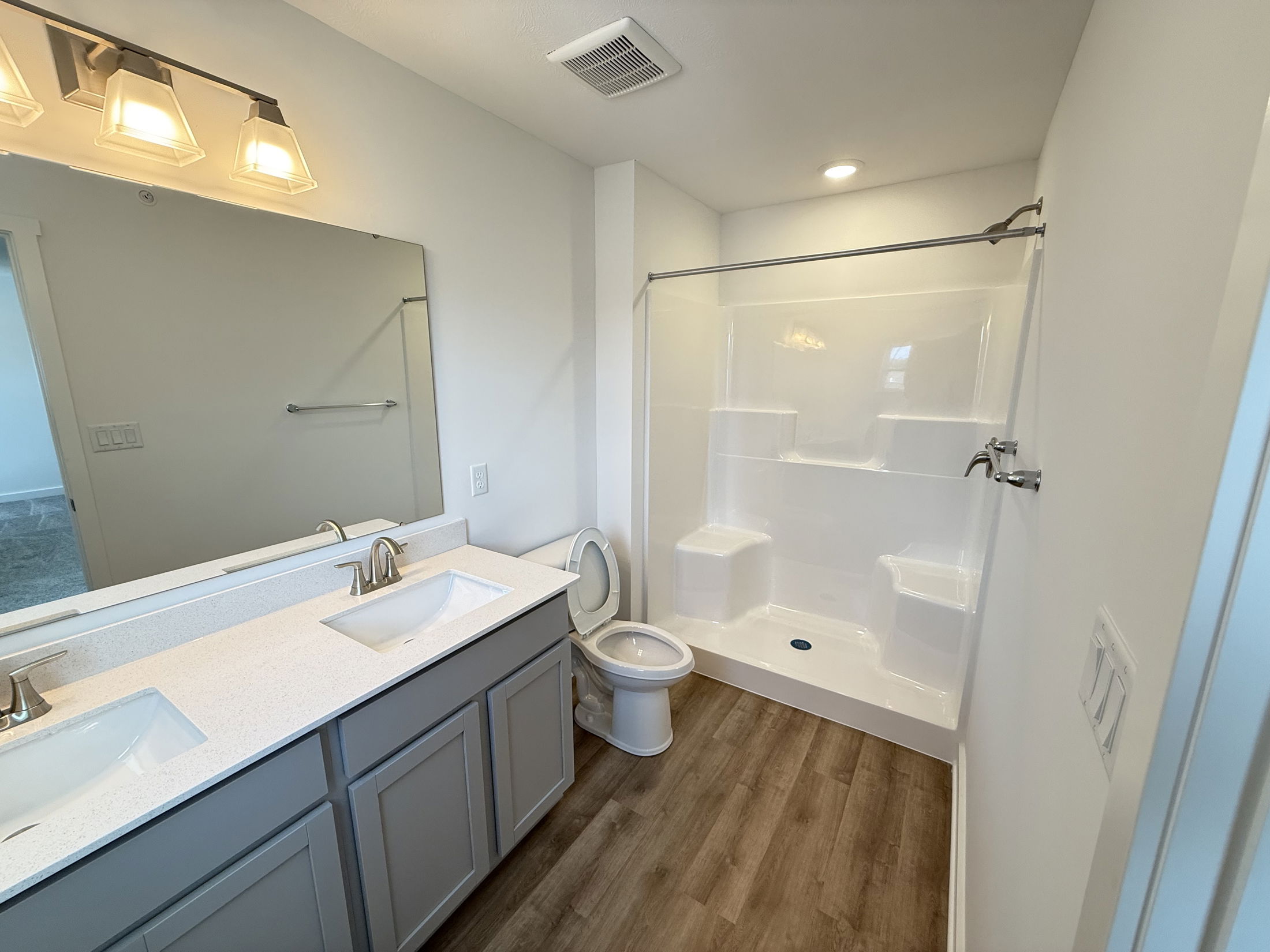 Modern bathroom with dual sinks, large mirror, toilet, and spacious shower featuring wood-style flooring.