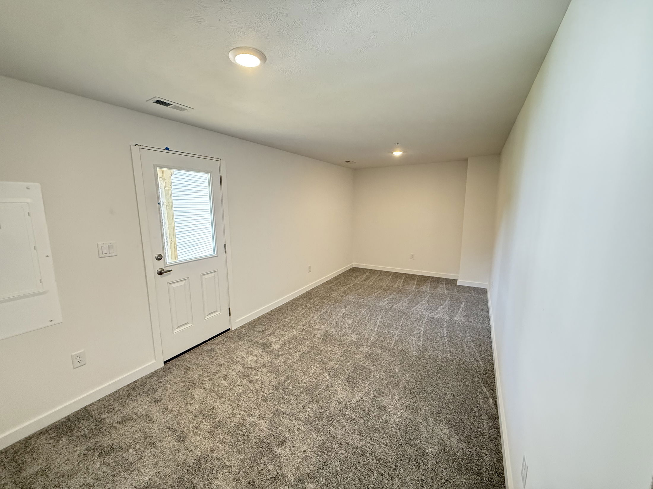 Wide-angle view of an empty room with plush gray carpet, white walls, recessed lighting, and a door with a small window.