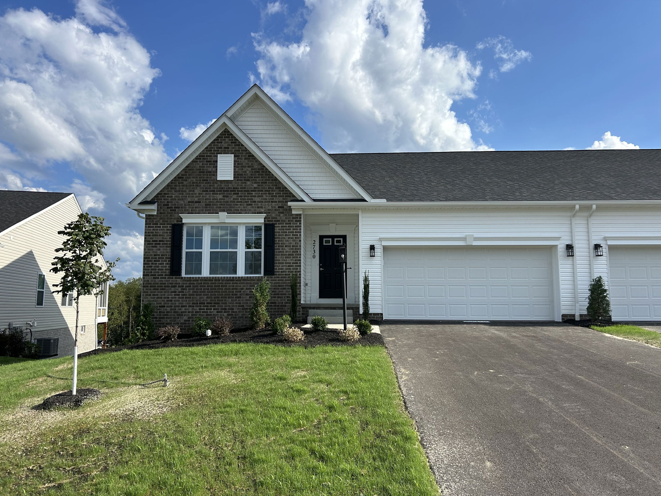 Modern suburban house with a brick facade, attached two-car garage, and freshly planted lawn under a partly cloudy blue sky.