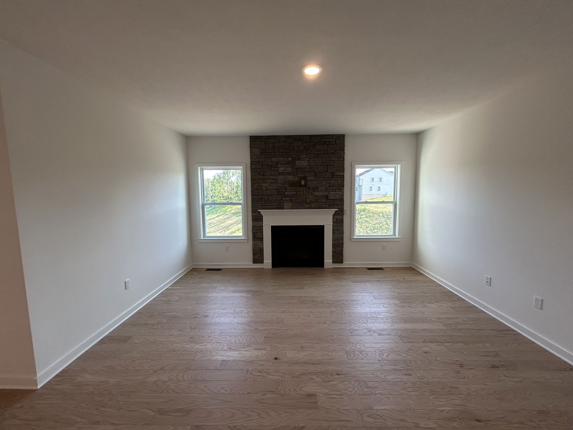 Spacious living room with wooden floors, a central stone fireplace, and windows providing ample natural light.