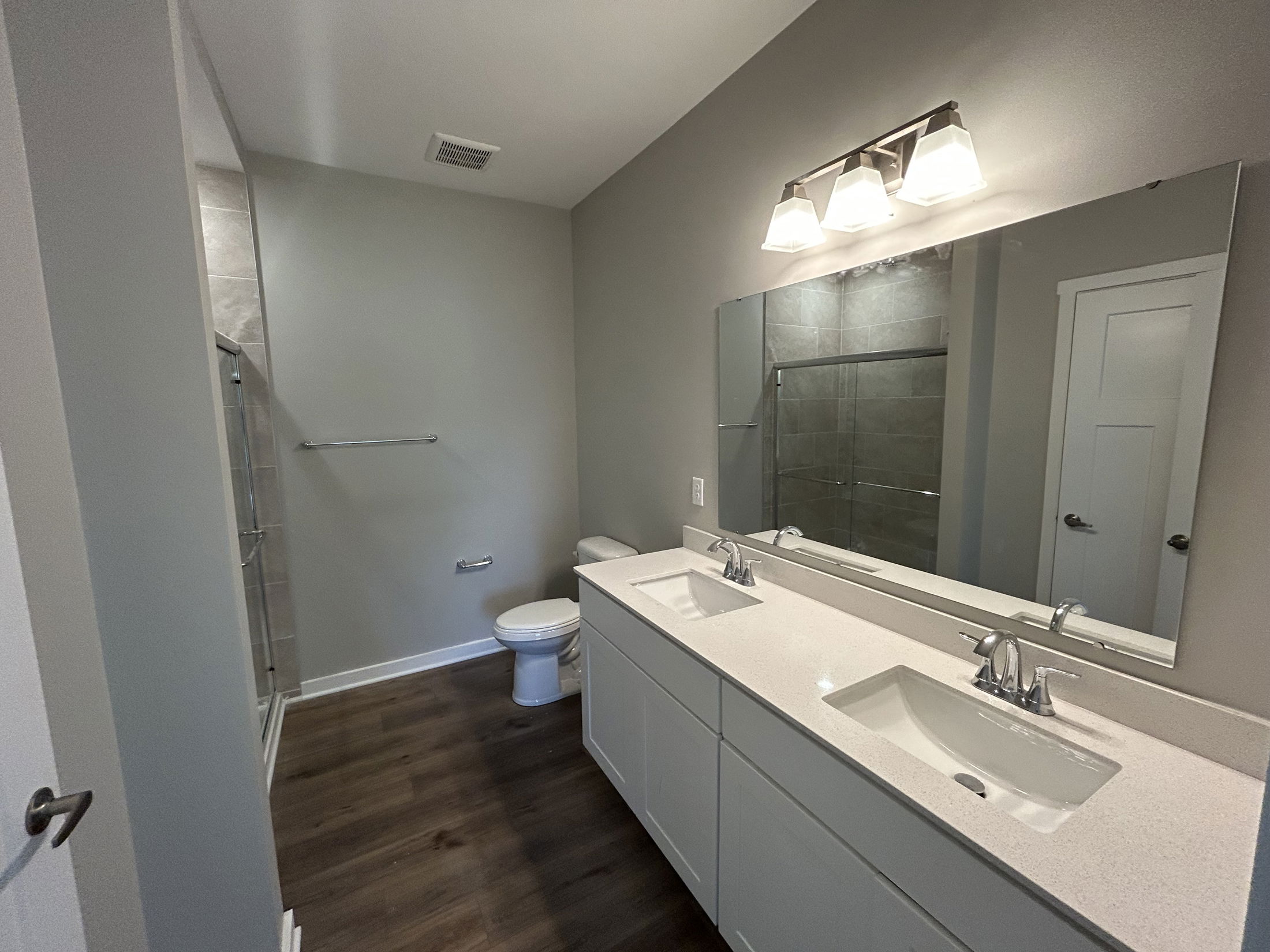 Modern bathroom with dual sinks, large mirror, glass-enclosed shower, and wooden flooring.