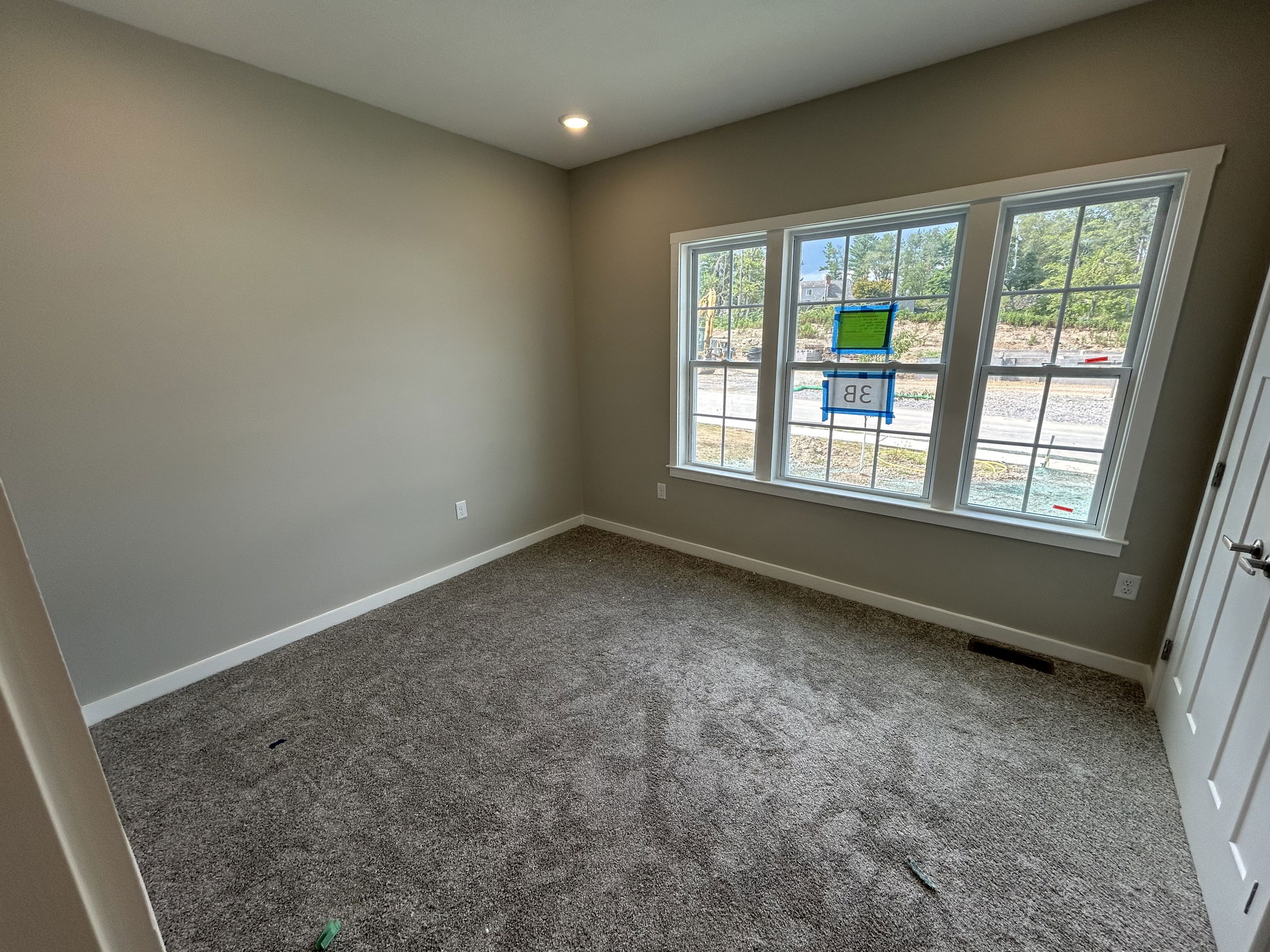 A newly painted, empty room with gray carpet and a large window overlooking a residential construction site.