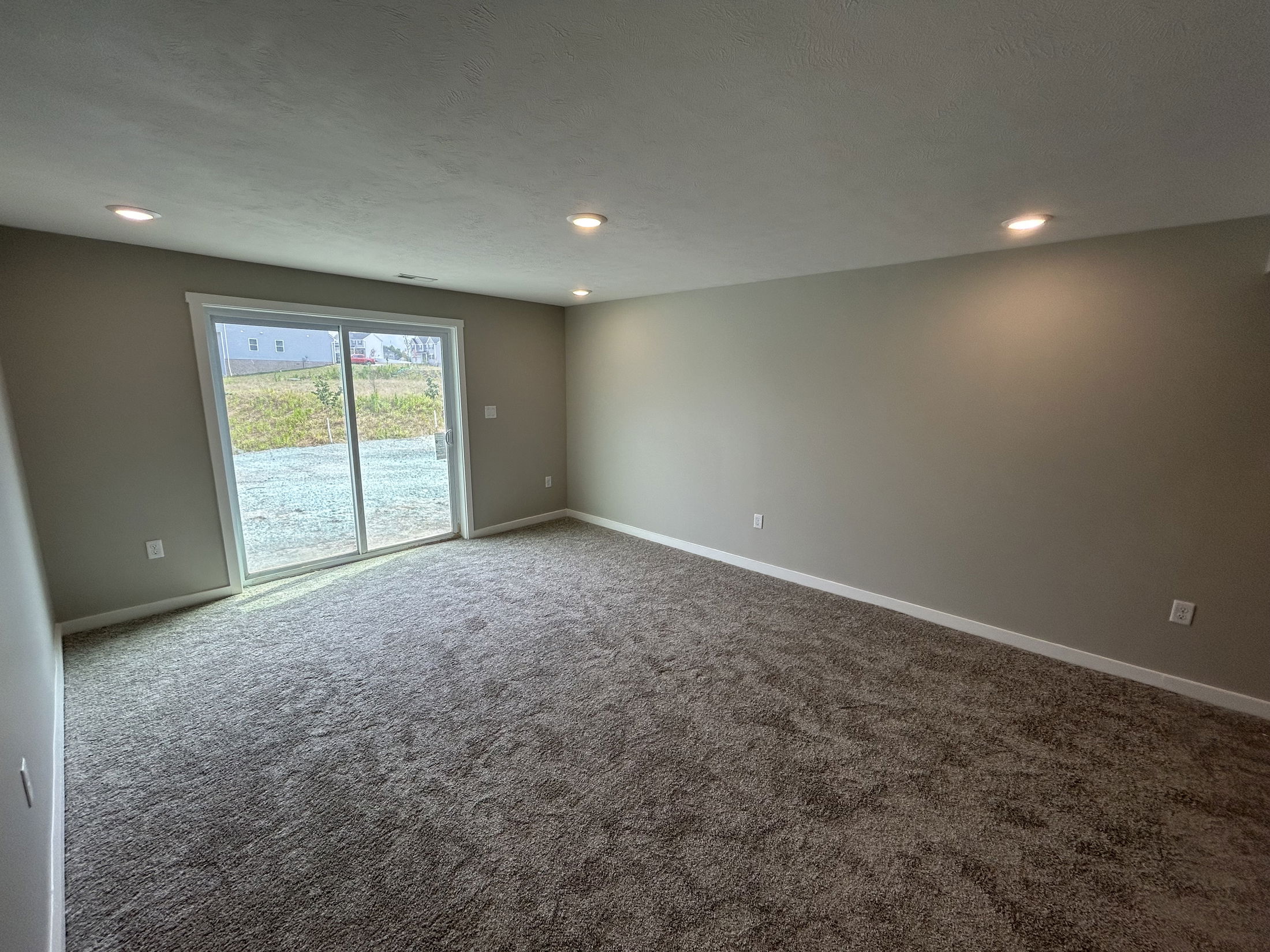 Empty gray-carpeted room with recessed lighting and a large sliding glass door leading outside.
