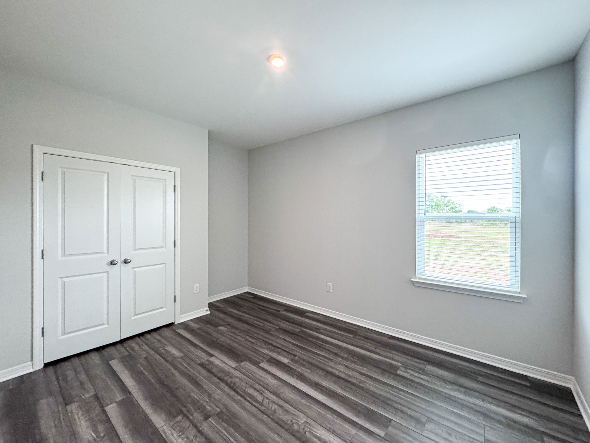 Empty modern bedroom interior with gray laminate flooring, white walls, double closet doors, and a window featuring white blinds.