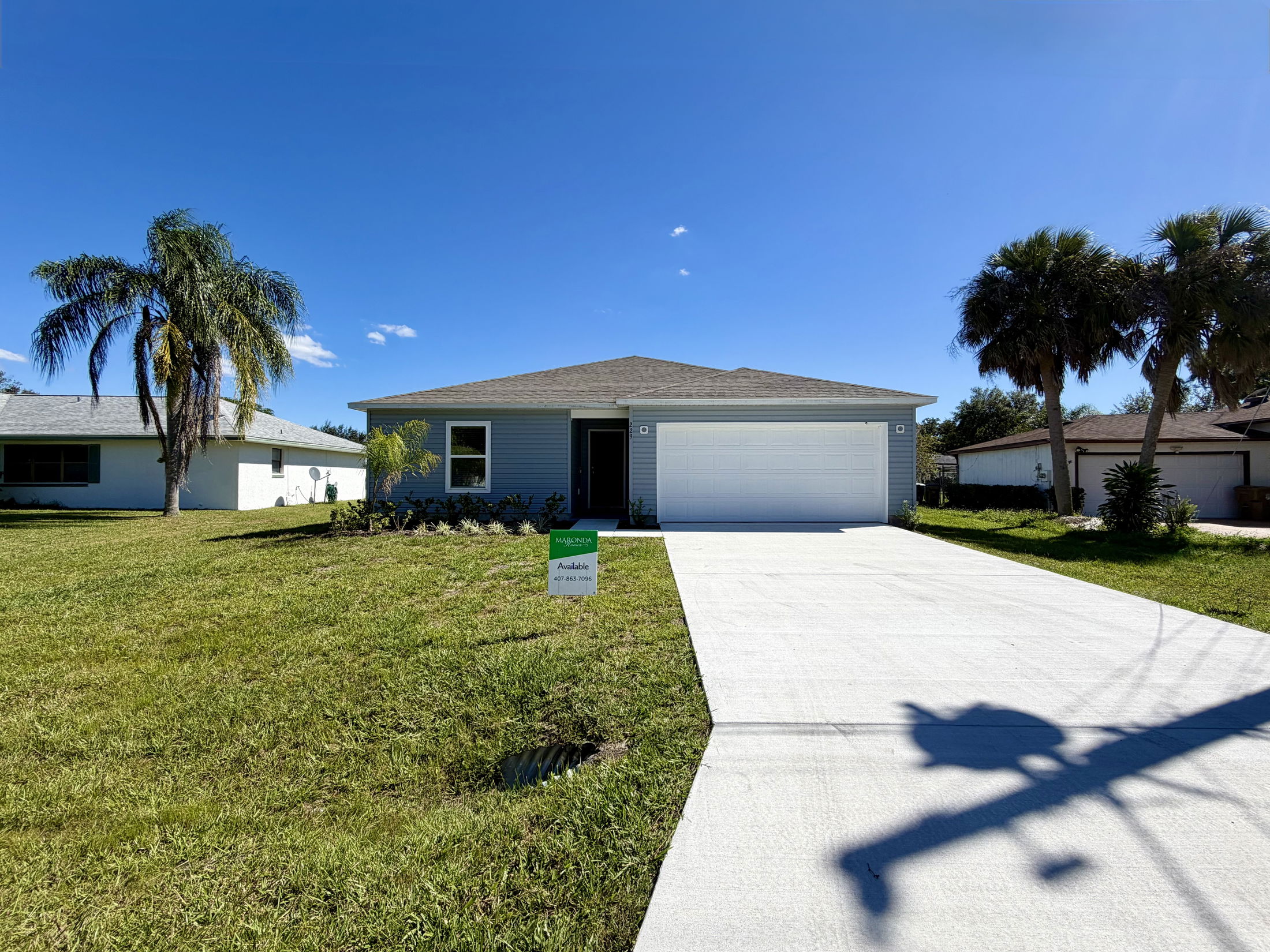 Newly constructed single-story home with a two-car garage, available for sale, featuring a spacious lawn and palm trees under a clear blue sky.