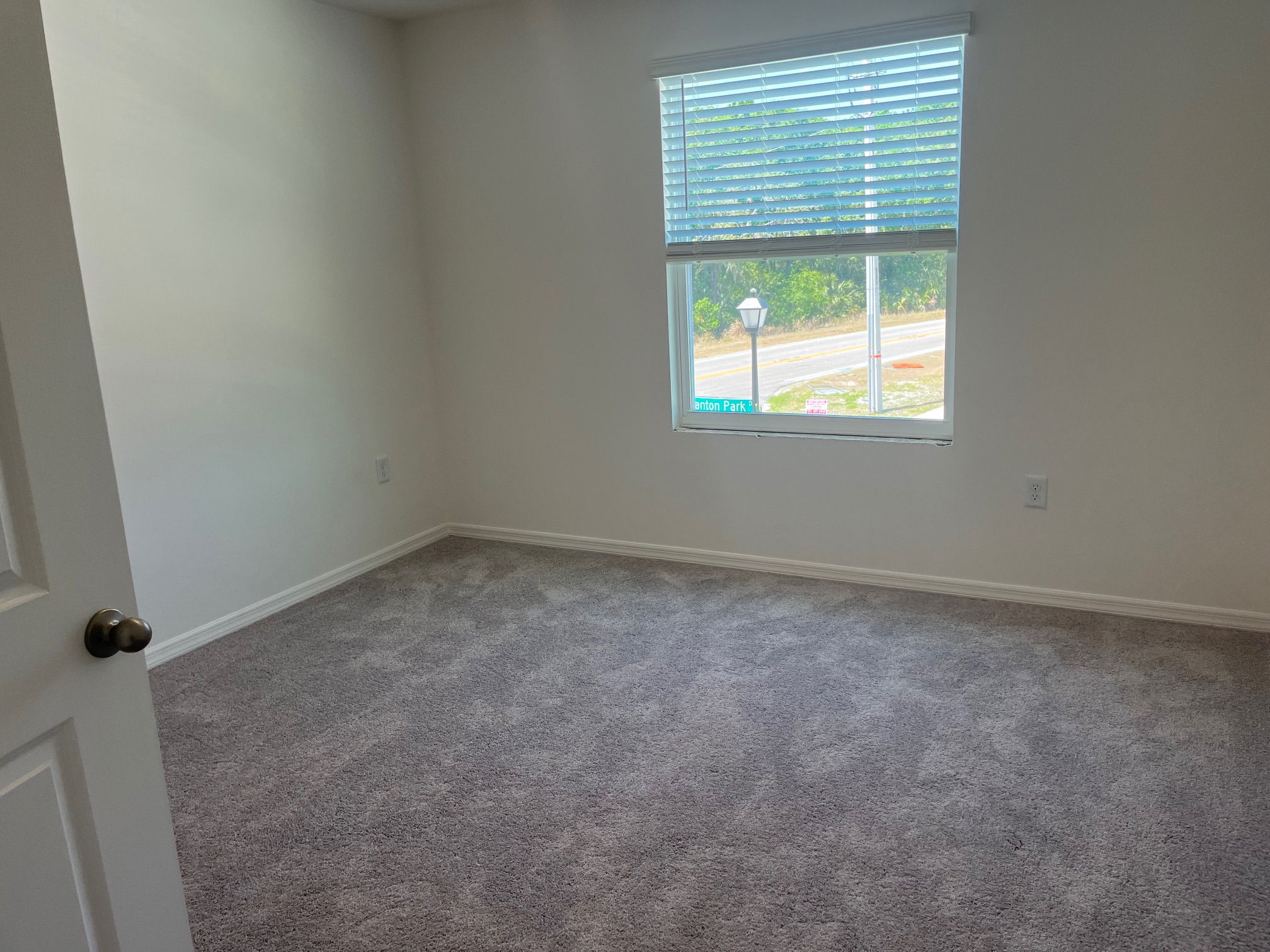 Empty room with gray carpet, white walls, and a window overlooking a street with greenery.