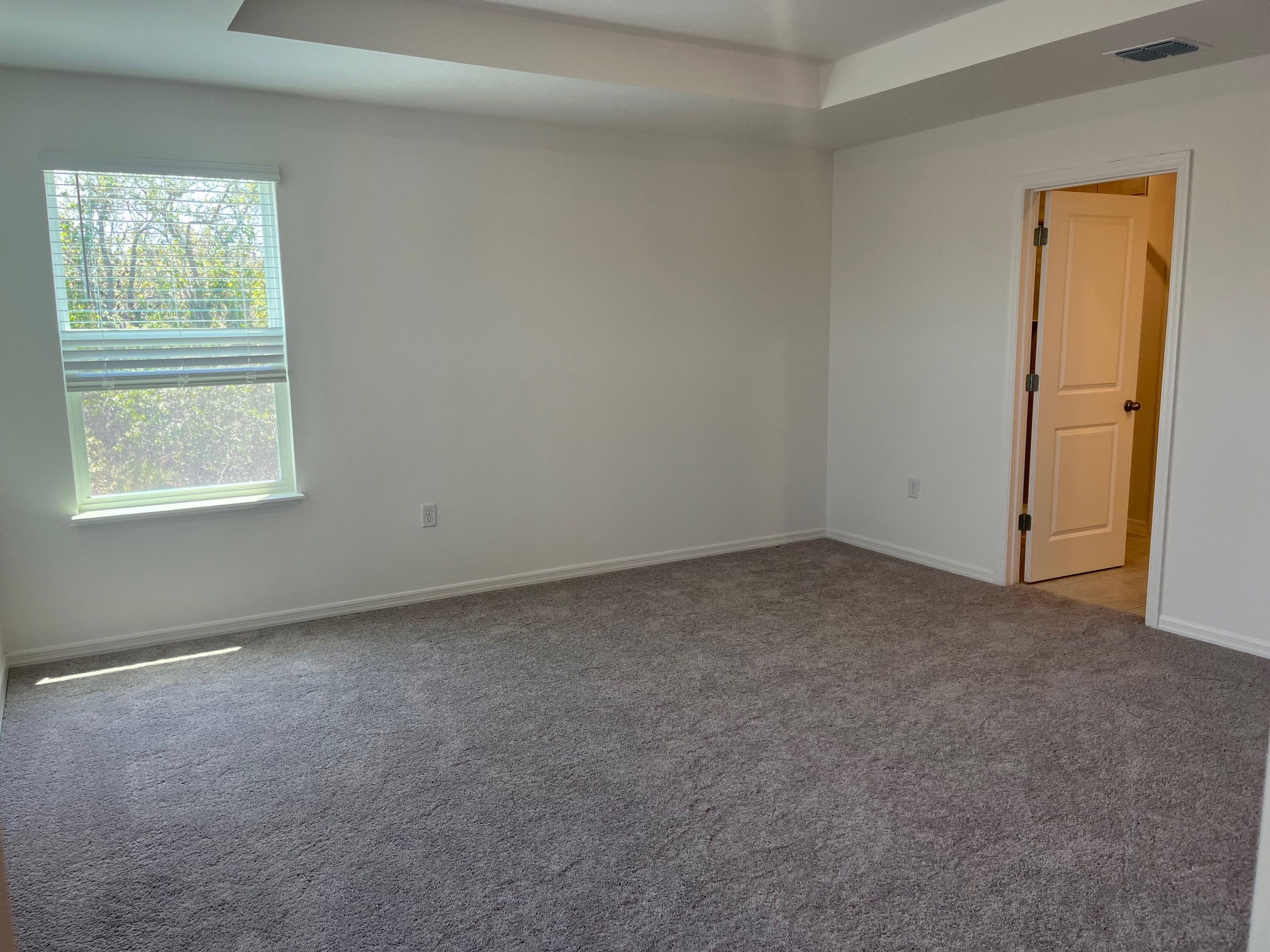 Spacious, empty carpeted room with a window and an open door leading to an adjacent area.