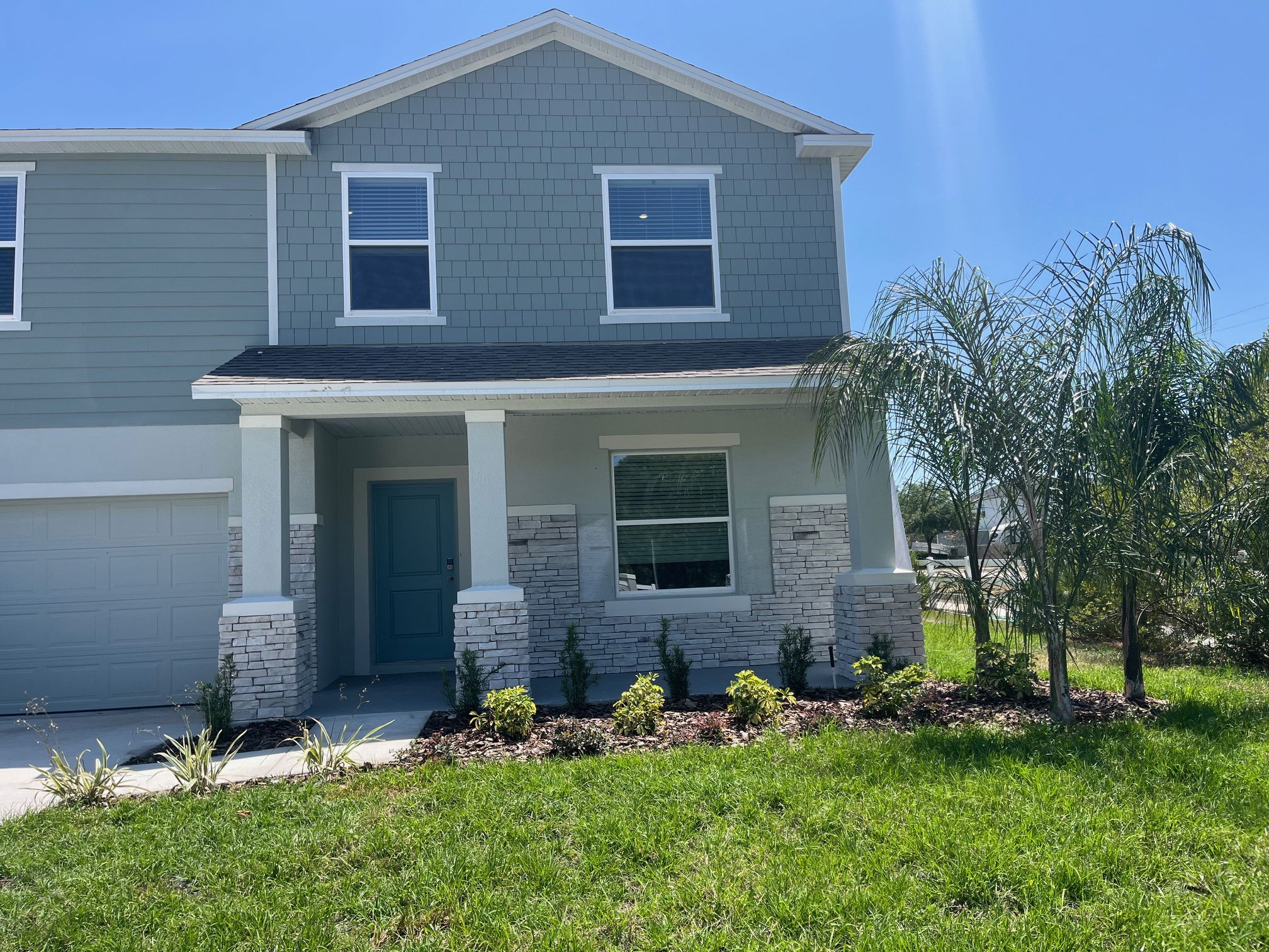 Modern two-story house with stone accents, blue siding, and a landscaped front yard under a clear blue sky.