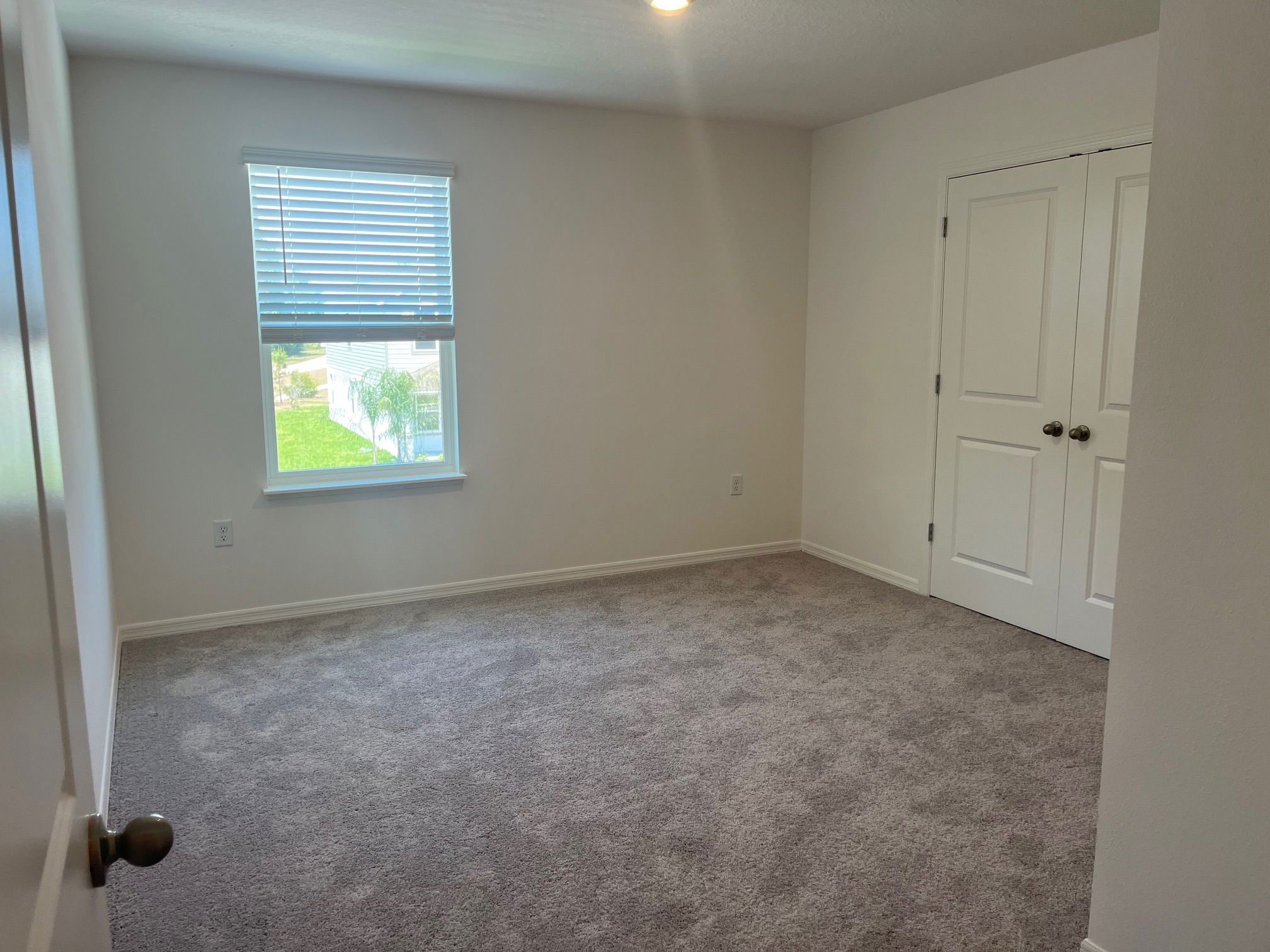 Empty carpeted room with a window featuring blinds and a closed double-door closet.