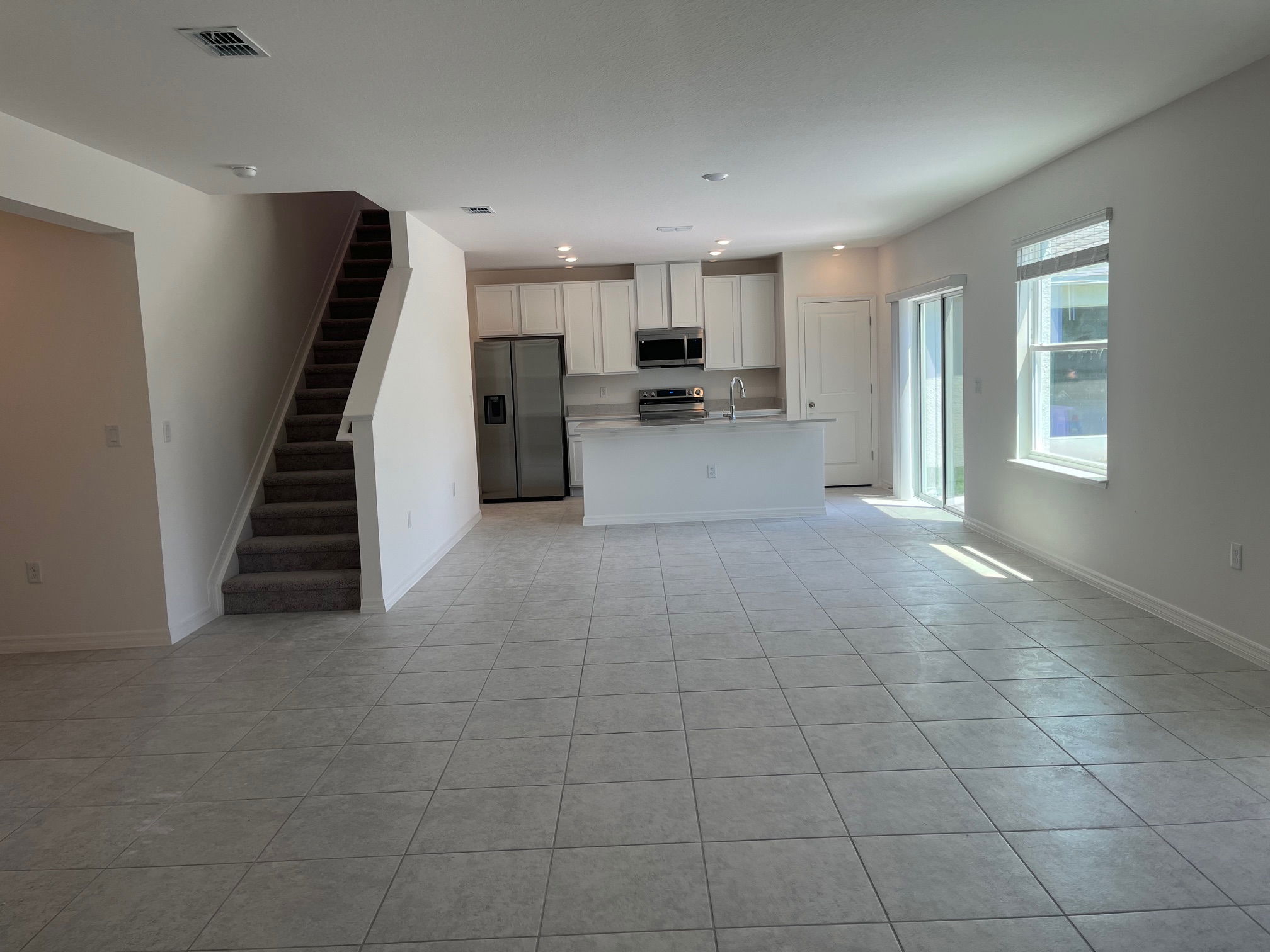 Spacious modern kitchen and living area with tiled floors, white cabinetry, and a staircase leading upstairs, illuminated by natural light.