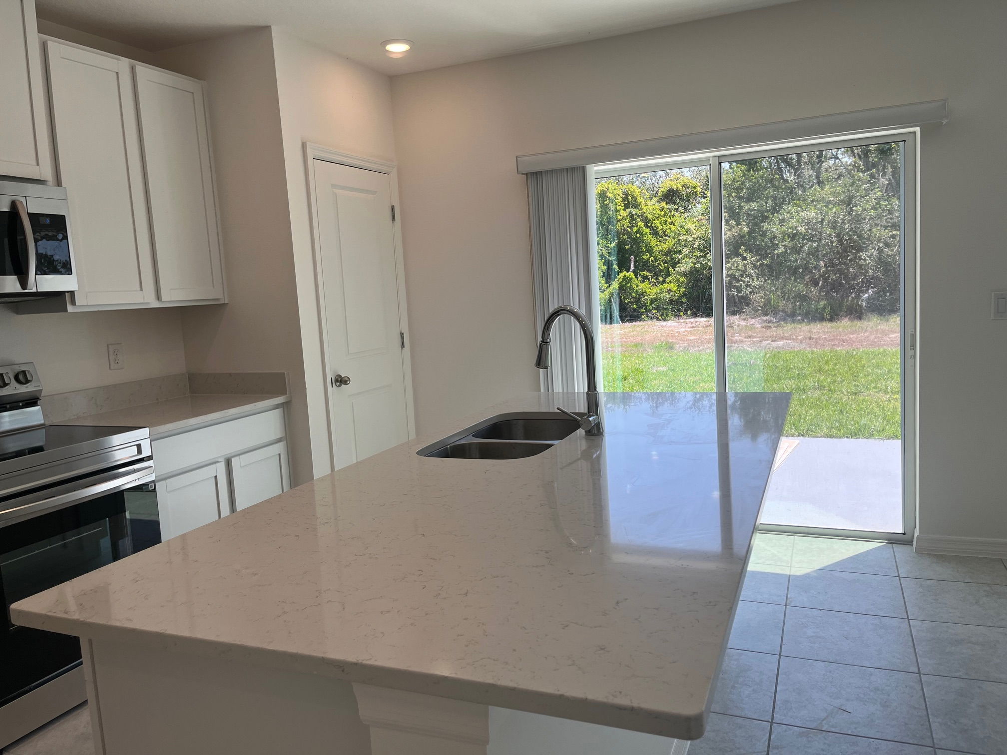 Modern kitchen with white cabinetry, stainless steel appliances, and a large quartz countertop island next to a sliding glass door overlooking a lush backyard.
