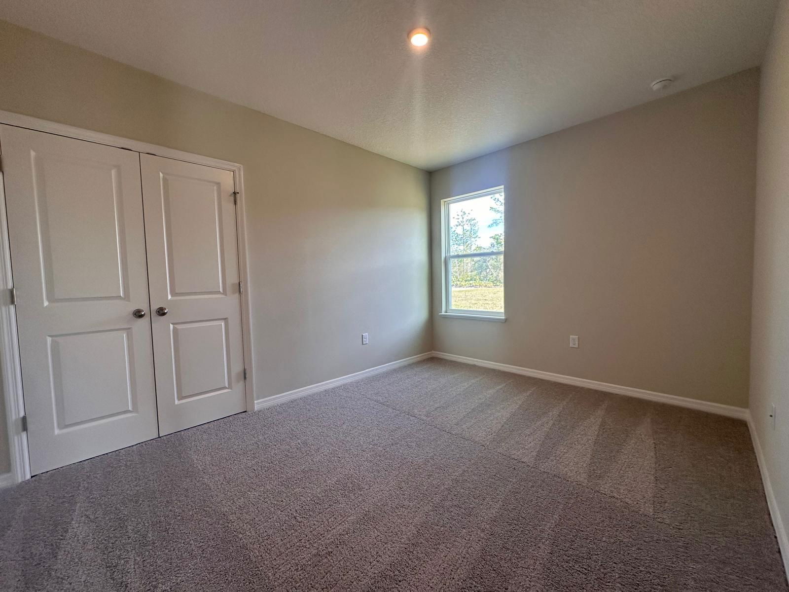 Empty beige carpeted room with a large window and double closet doors, perfect for a bedroom or office.