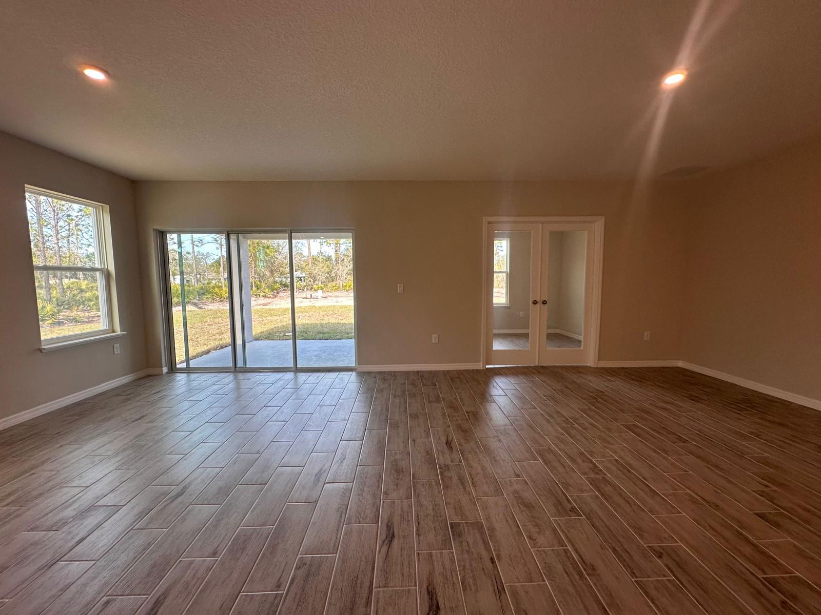 Spacious, empty living room with wood-look tile flooring, large windows, and sliding glass doors opening to a patio and greenery.