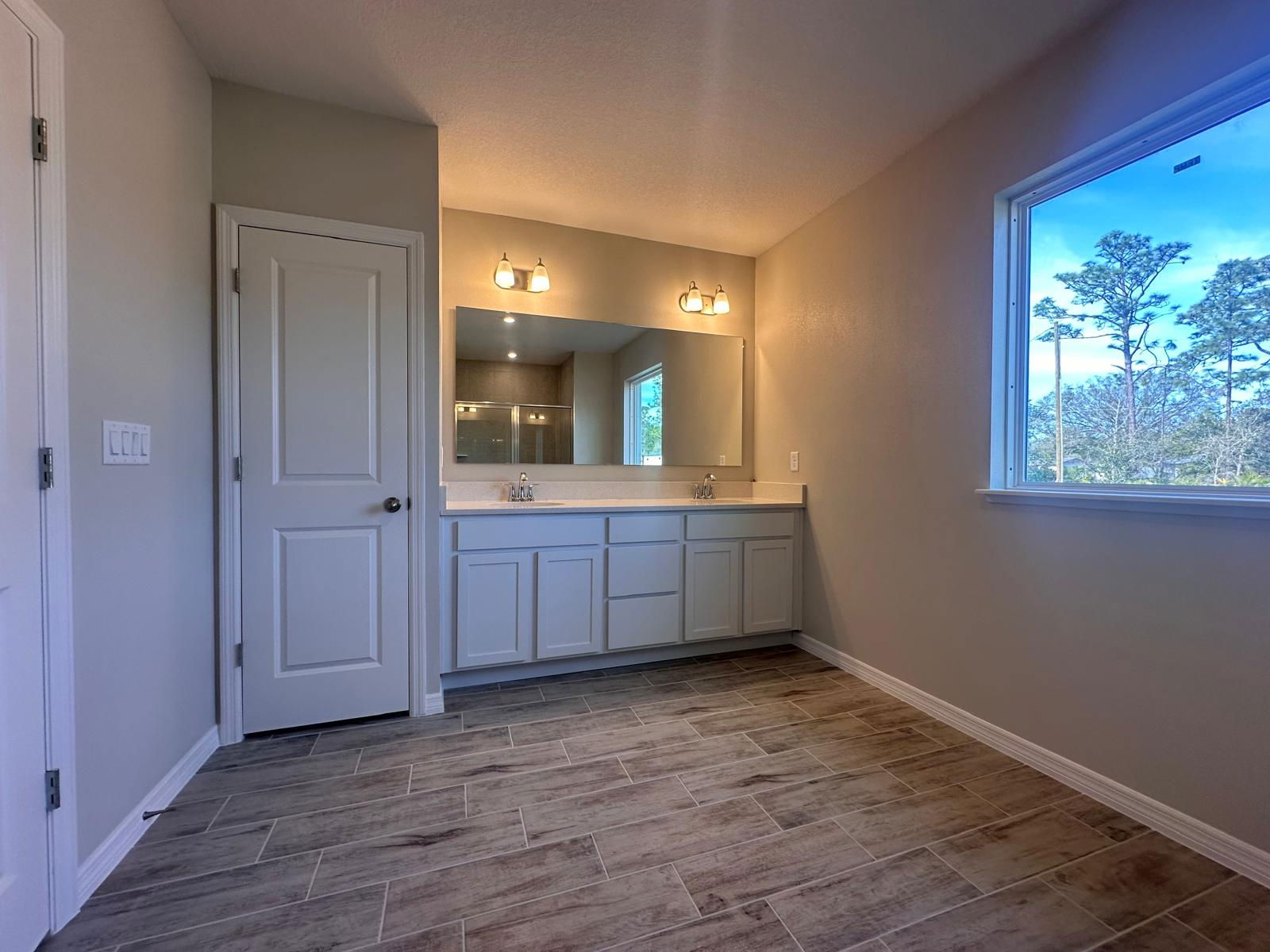 Modern bathroom with dual lights, a large mirror, white cabinetry, and wood-style tile flooring, featuring a scenic window view.