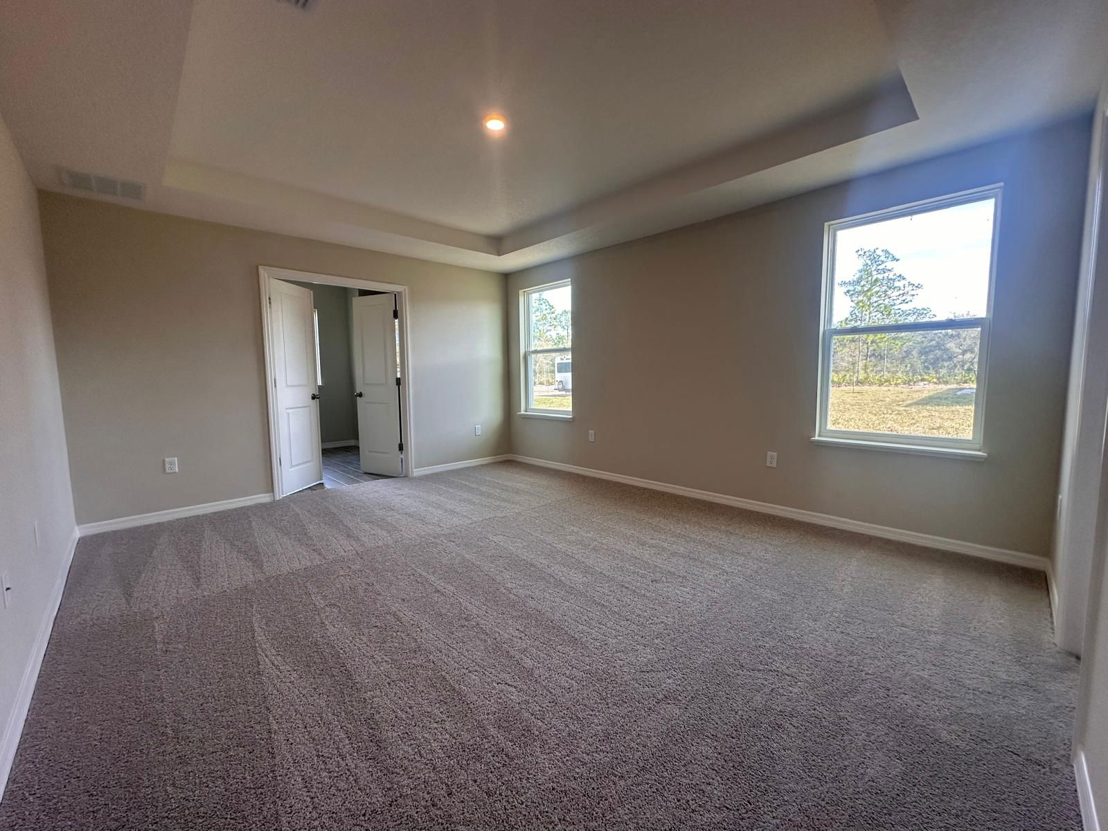 Spacious, empty beige bedroom with carpet flooring, large windows, and an open double door leading to another room.