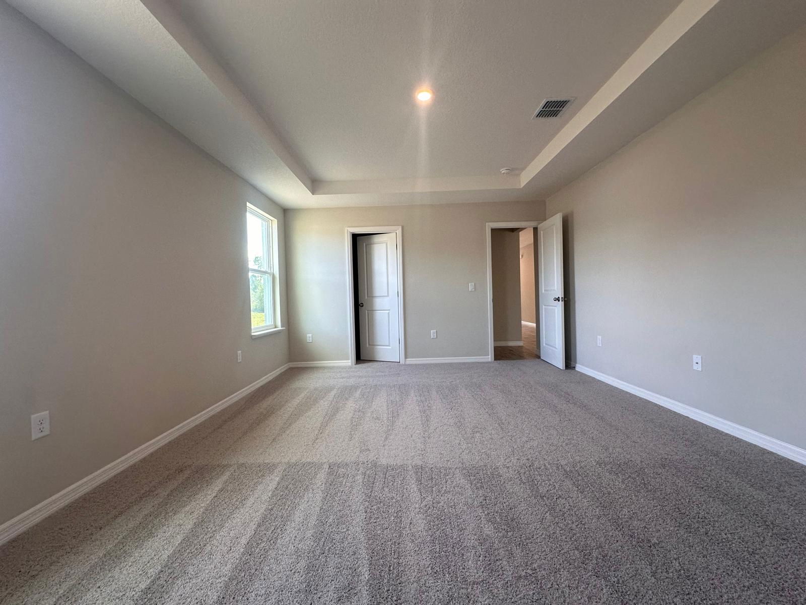 Spacious empty bedroom with beige carpeting, white doors, and a large window providing natural light.
