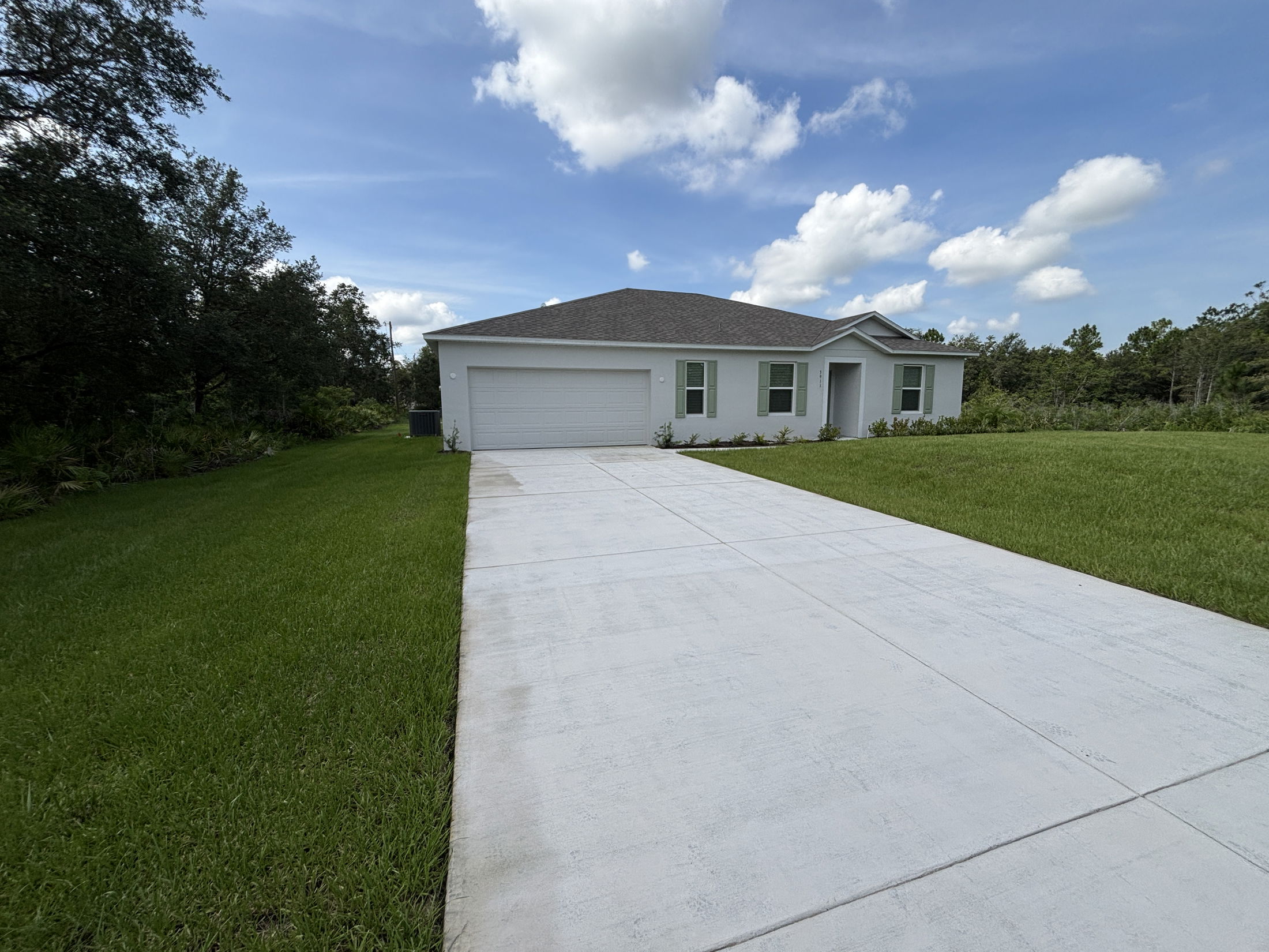 Newly built single-story white house with a gray roof, large driveway, and green lawn under a blue sky.