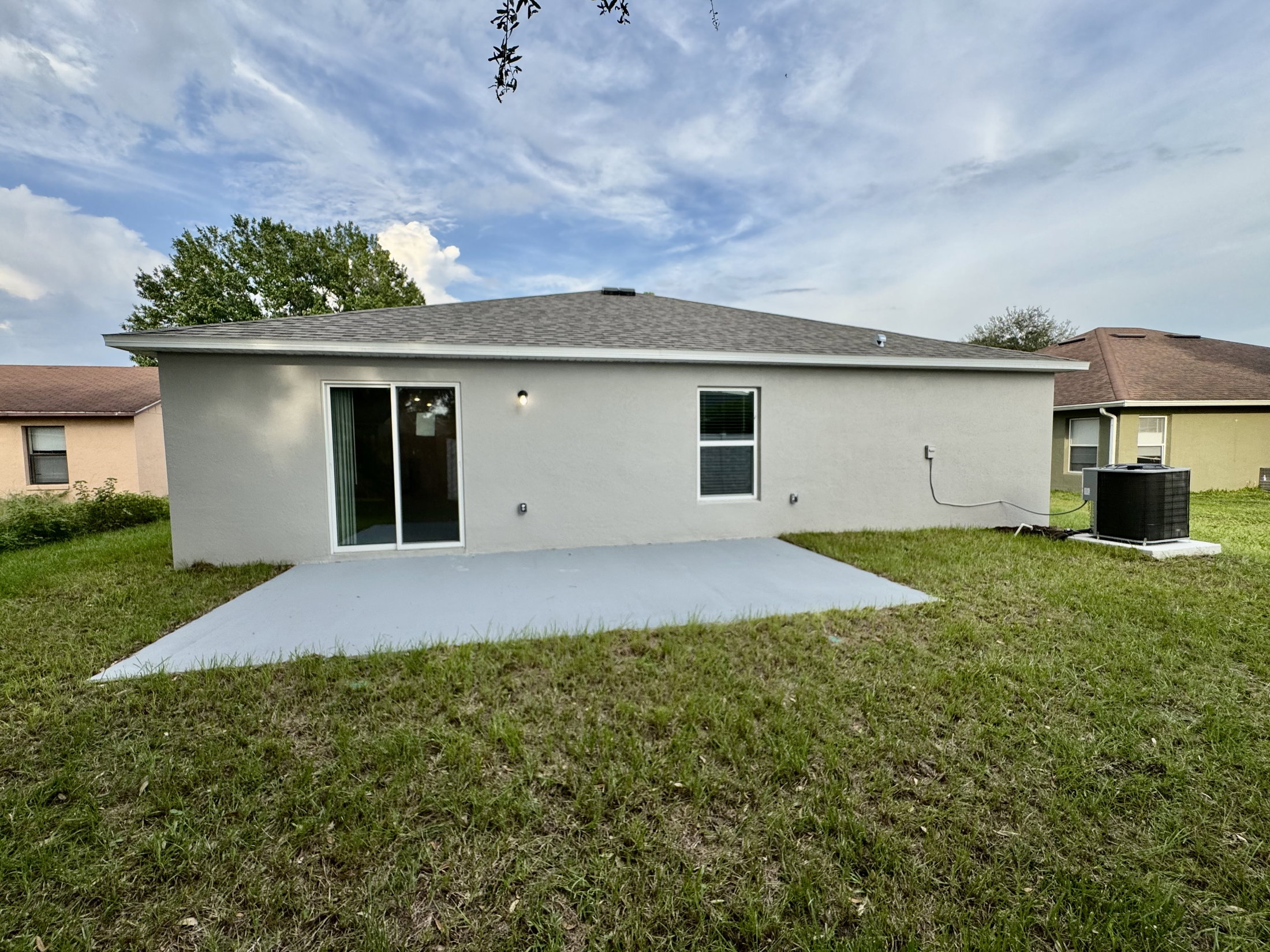 Back view of a single-story house with a concrete patio, surrounded by grass and trees.
