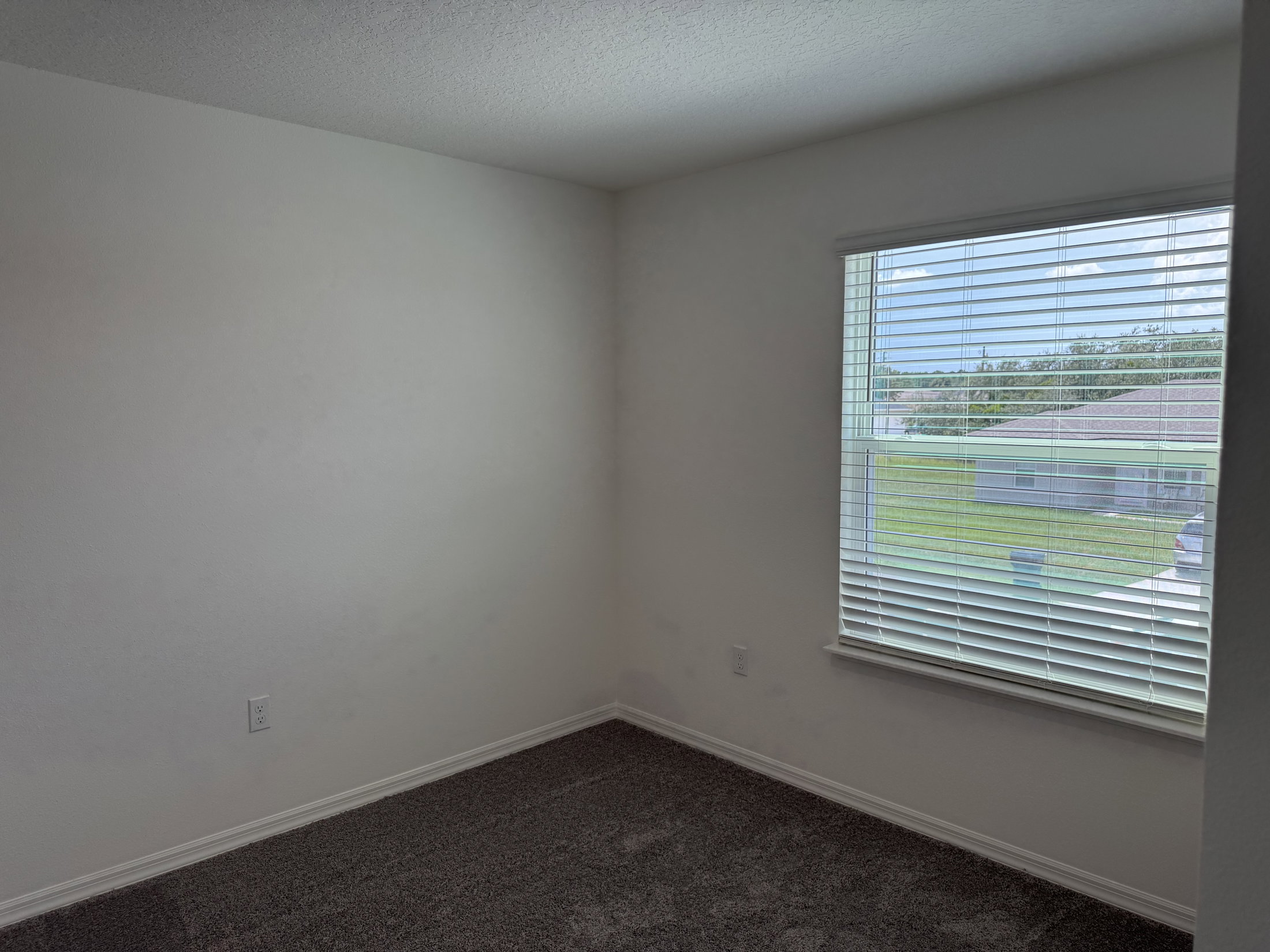 A vacant room with gray carpet, white walls, and a window with blinds letting in natural light.