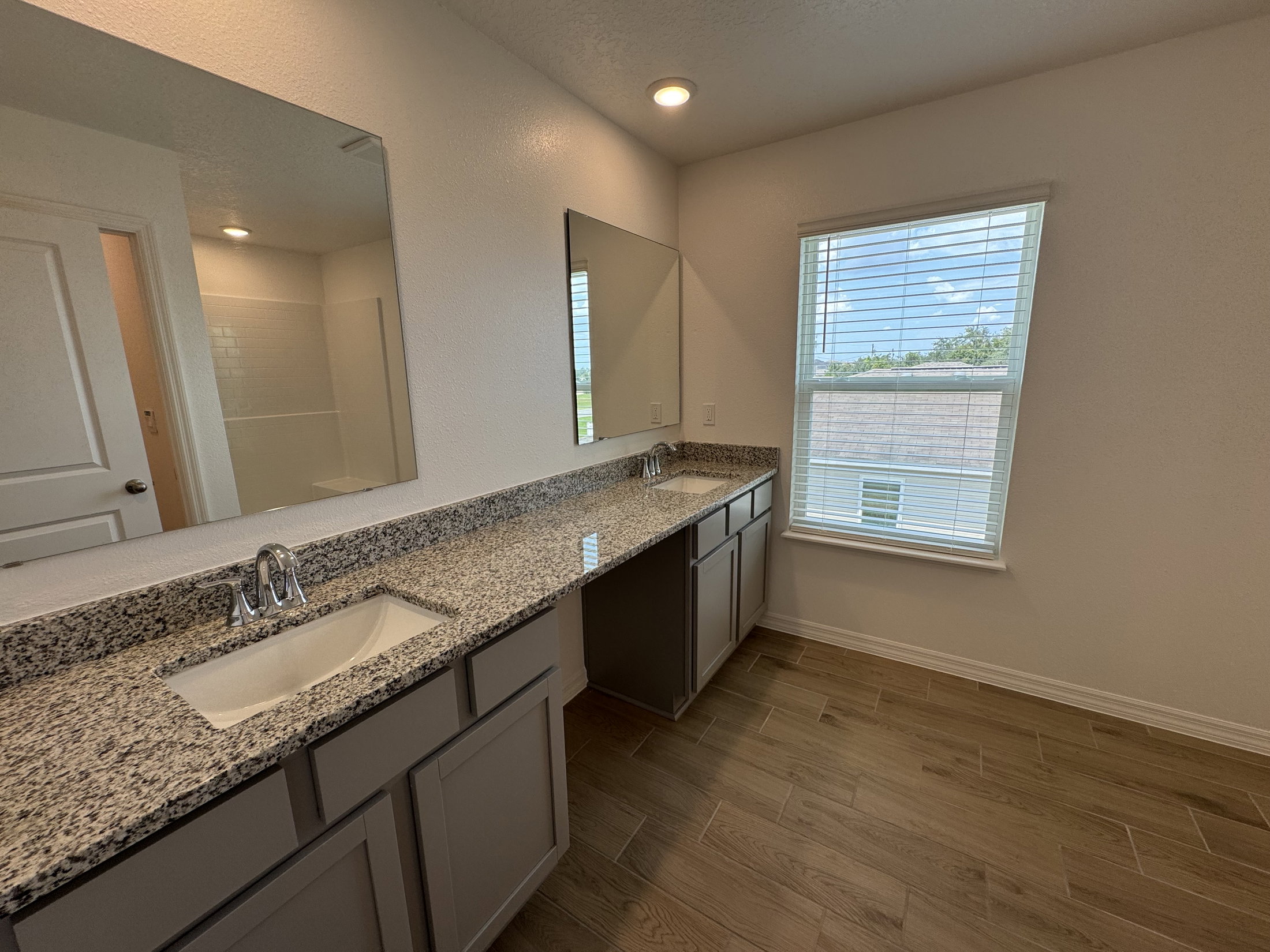 A clean, modern bathroom with a double vanity, granite countertops, and a window with blinds.