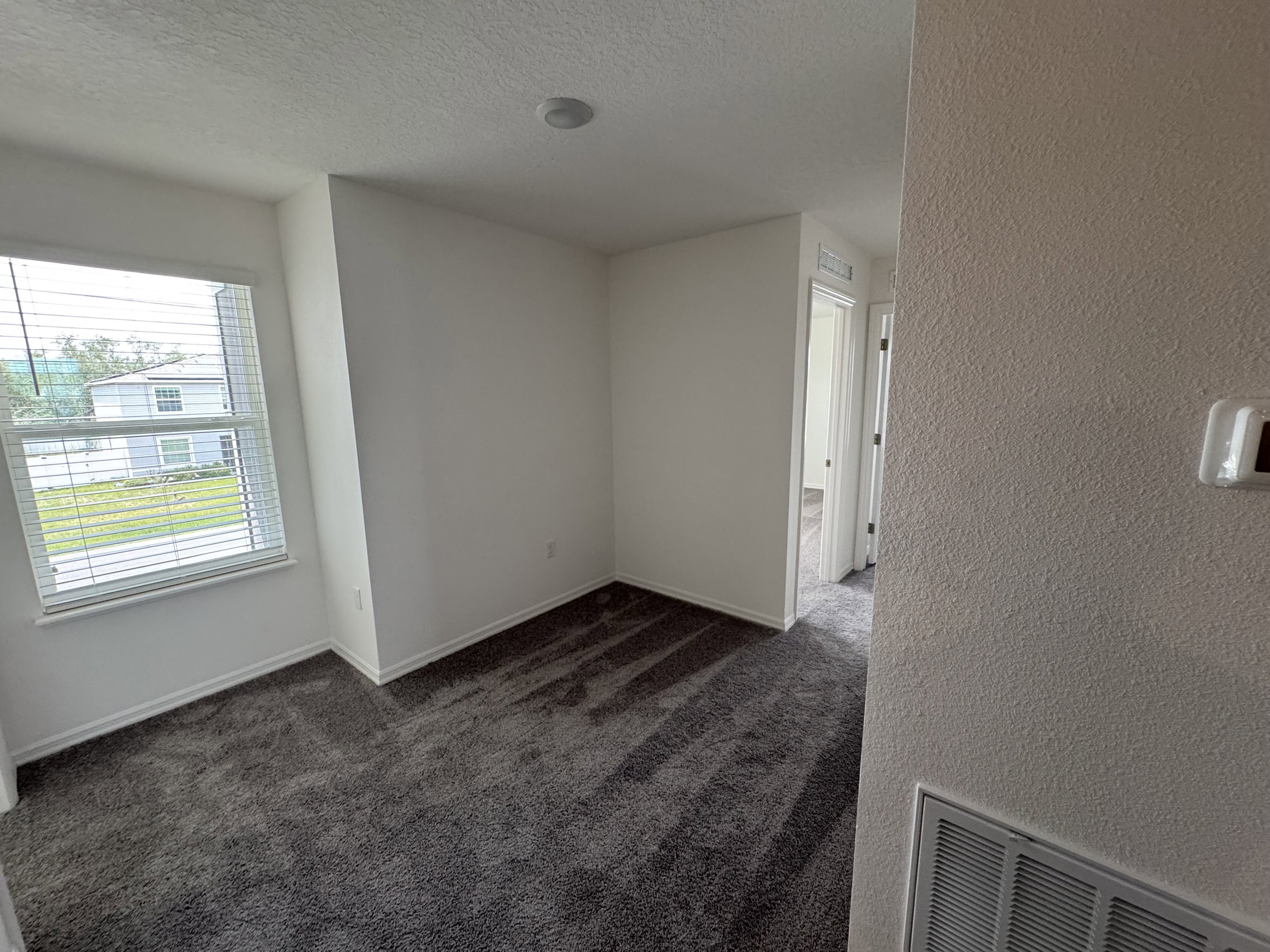 Empty room with light-colored walls, a window with blinds, and plush gray carpet.