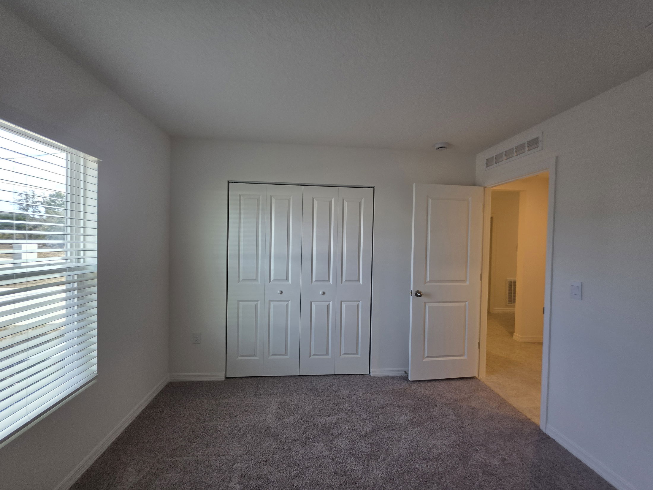 Spacious and bright bedroom interior featuring white walls, a window with blinds, and double closet doors.