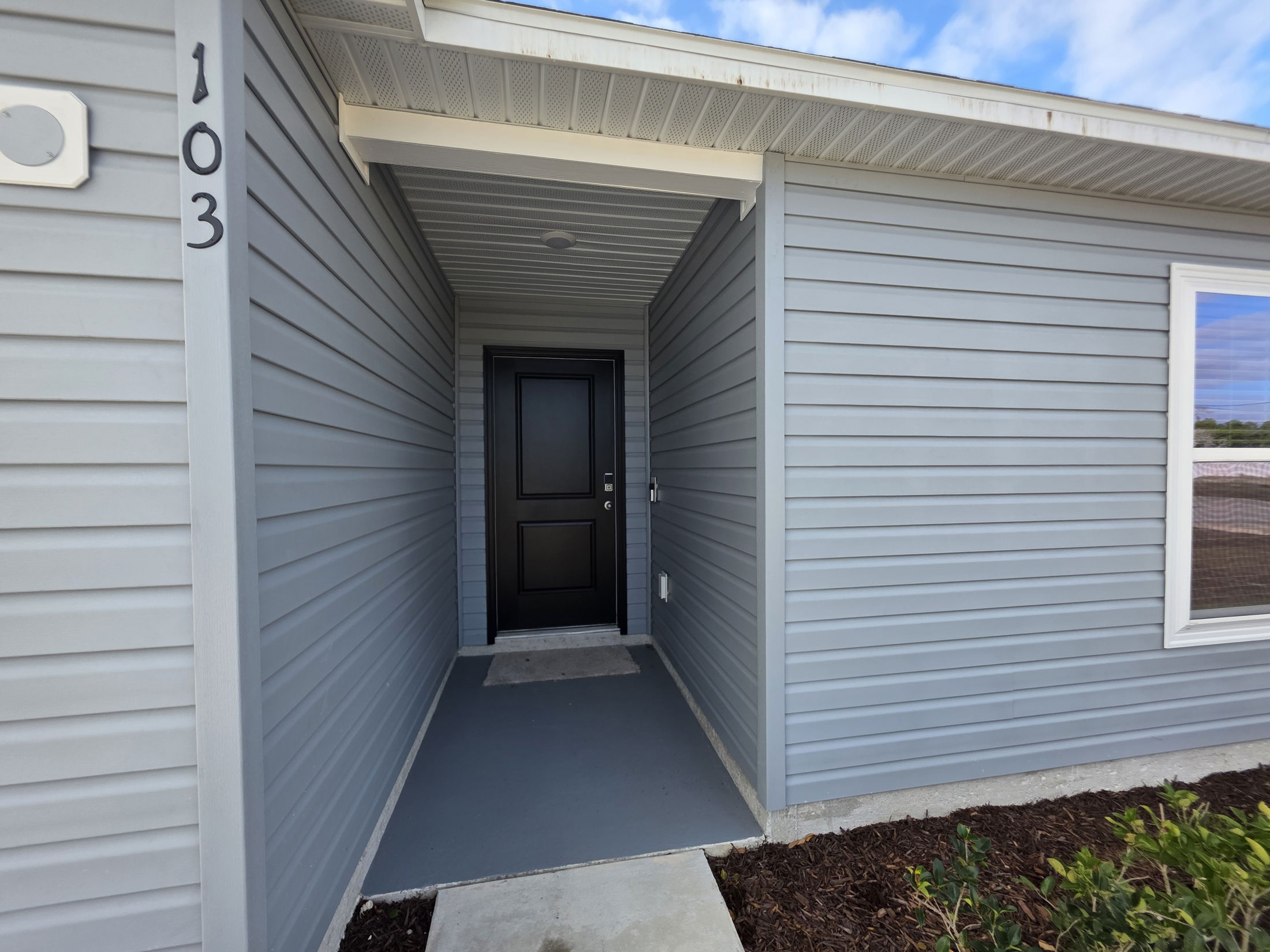 Exterior view of a modern home entrance with a black door and house number 103 on a gray siding background.