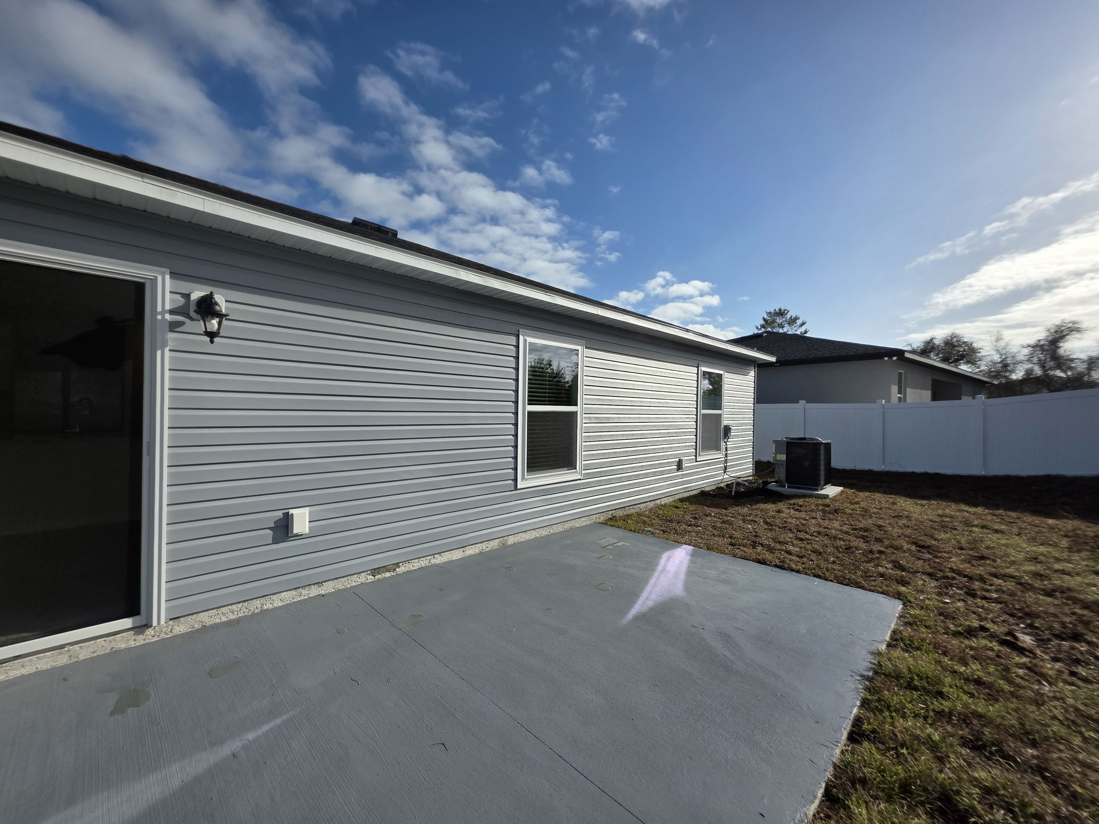 A newly constructed home exterior showcasing grey siding, a patio area, and a shaded backyard under a clear blue sky.