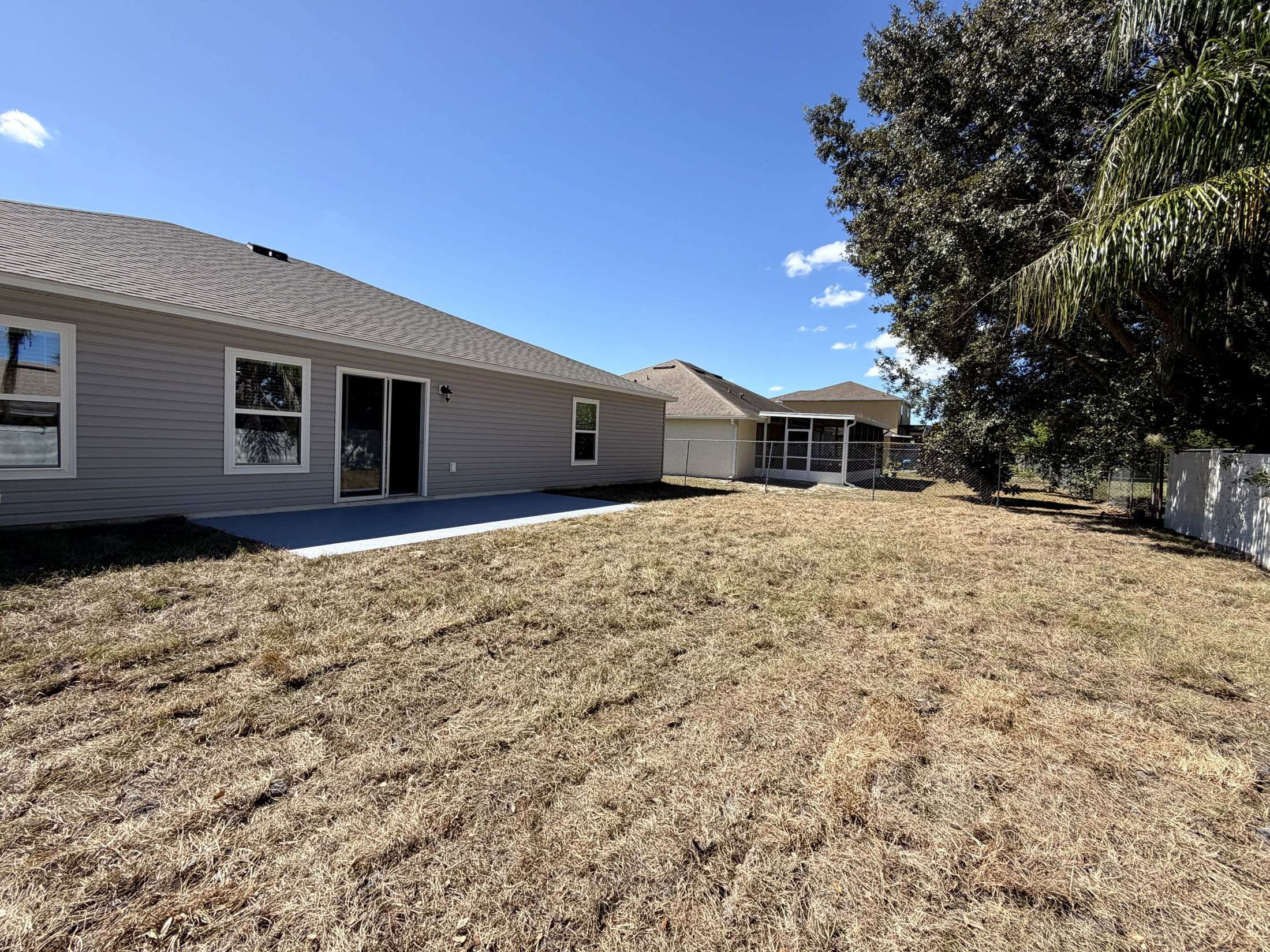 Backyard view of a suburban home with a spacious dry grass lawn, patio, and large tree under a clear blue sky.