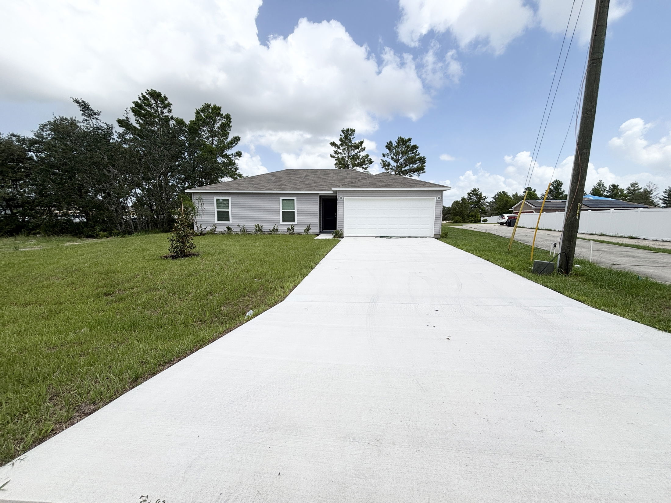 Suburban single-story house with a two-car garage, expansive driveway, and lush green lawn under a partly cloudy sky.