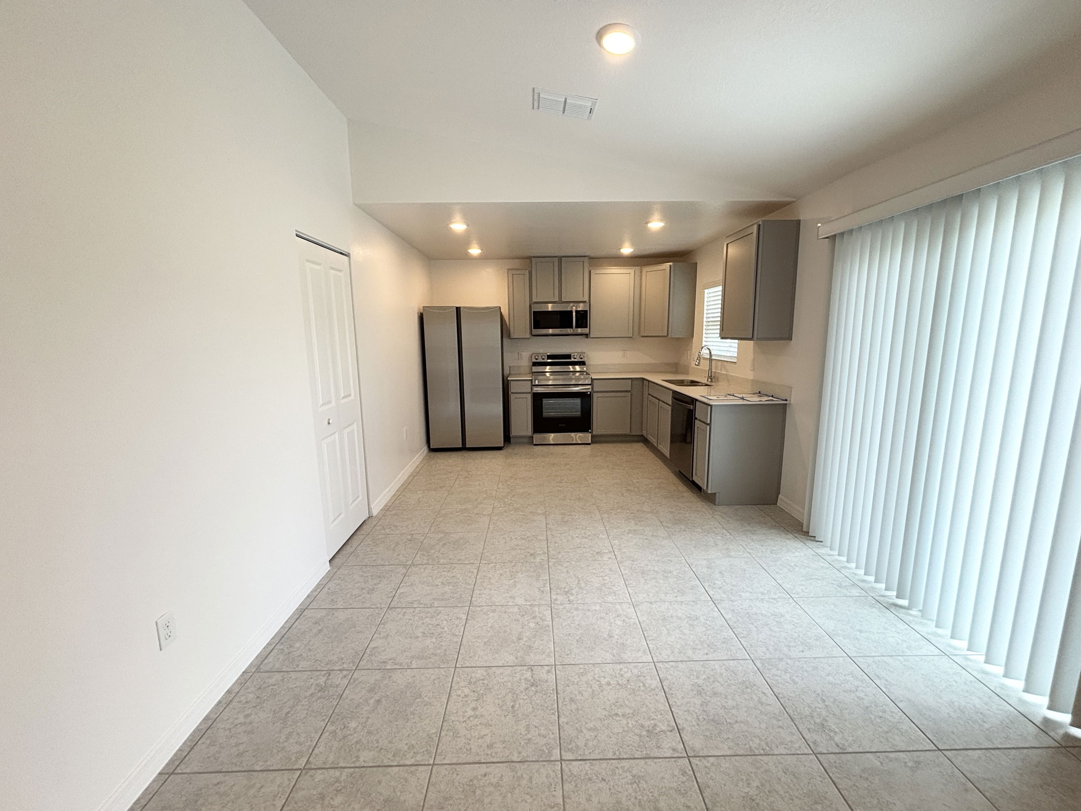 Open-concept kitchen with modern gray cabinets, stainless steel appliances, and large tile flooring, featuring natural light from sliding glass doors with vertical blinds.