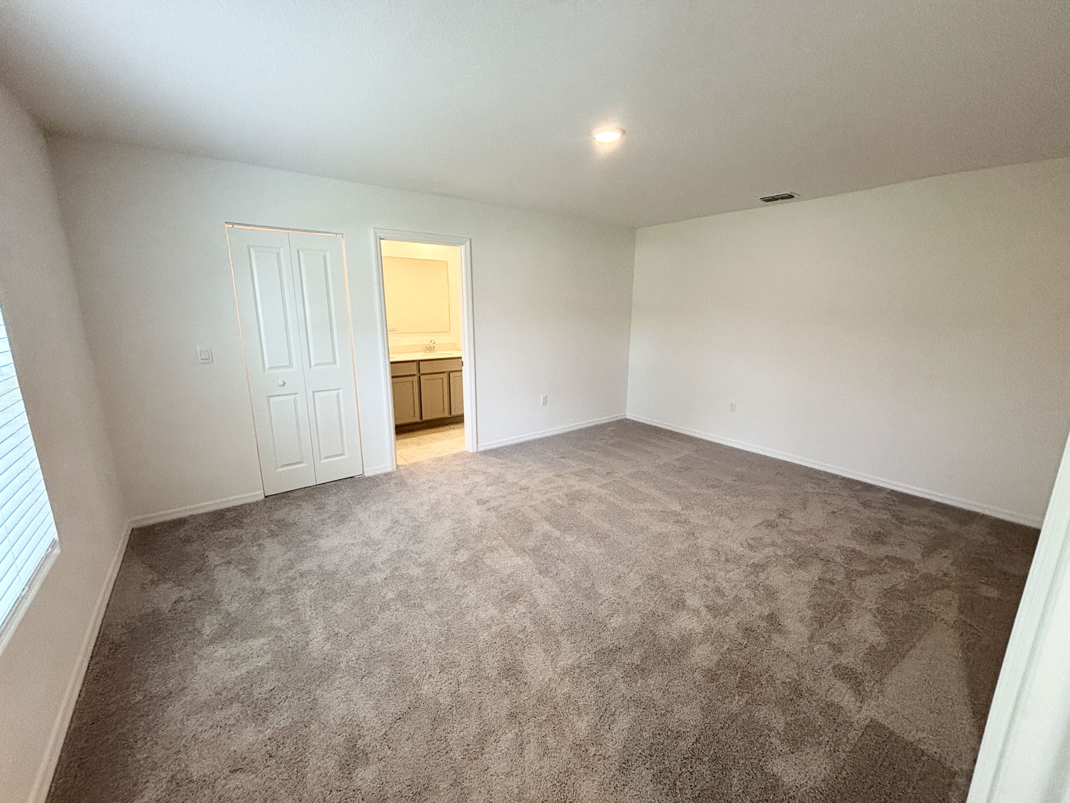 Empty carpeted bedroom with white walls, a window, a closet, and an open door leading to a bathroom.