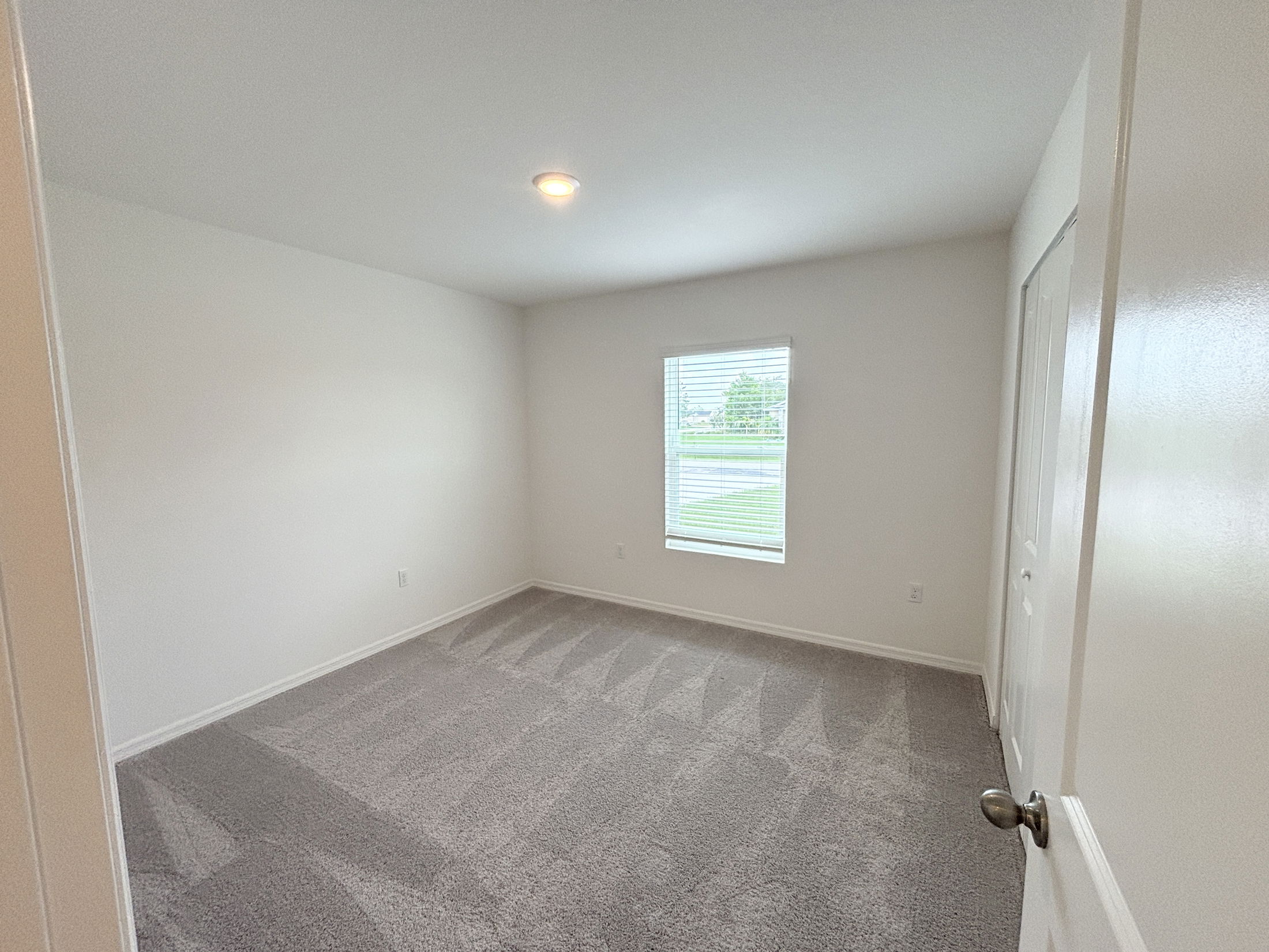 Empty bedroom with clean gray carpet, white walls, a window with blinds, and a closed door.