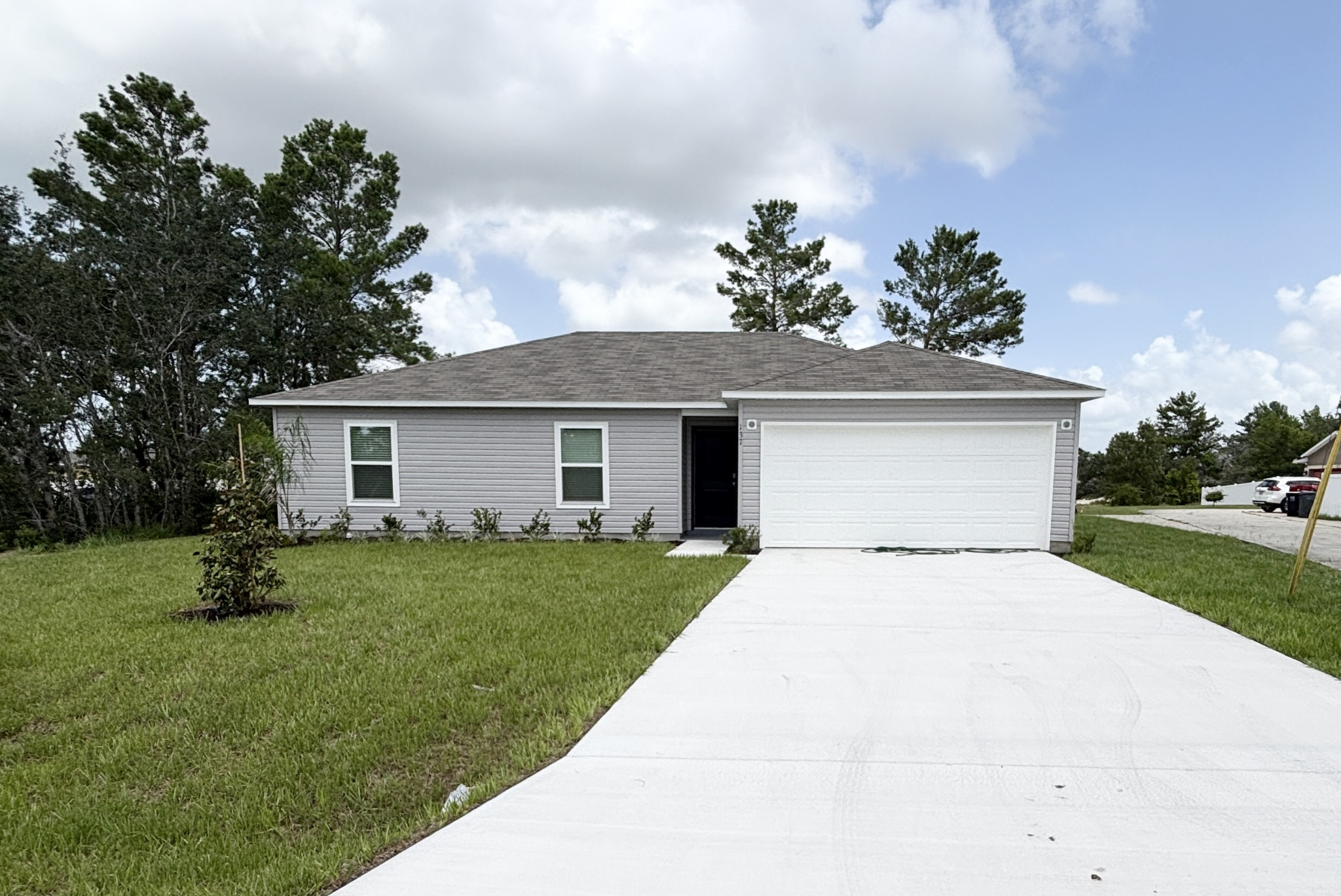 Modern single-story gray house with white garage on a freshly paved driveway, surrounded by a green lawn and trees.
