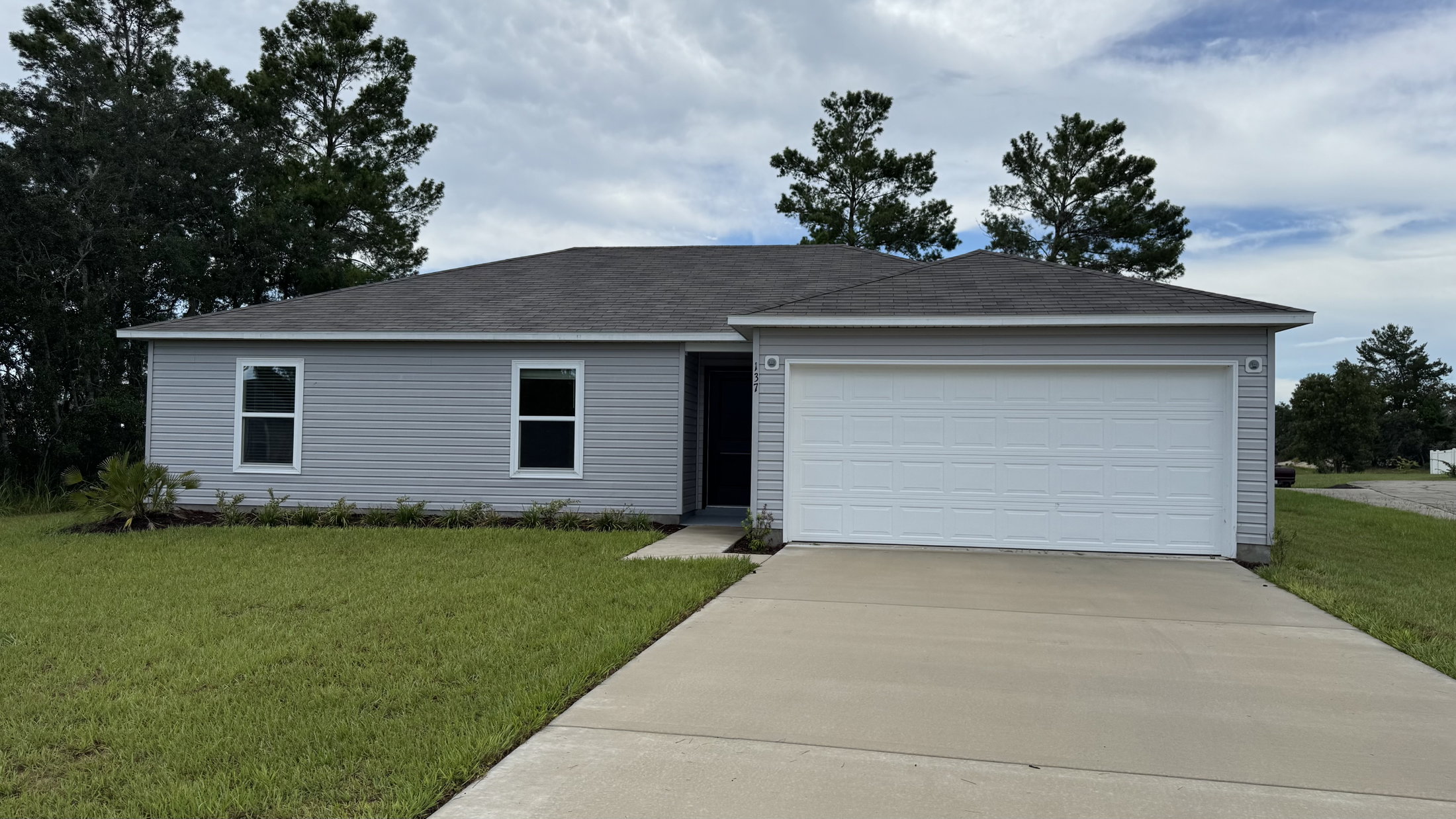 Single-story house with gray siding, a front porch, and a white garage door, surrounded by green grass and trees.