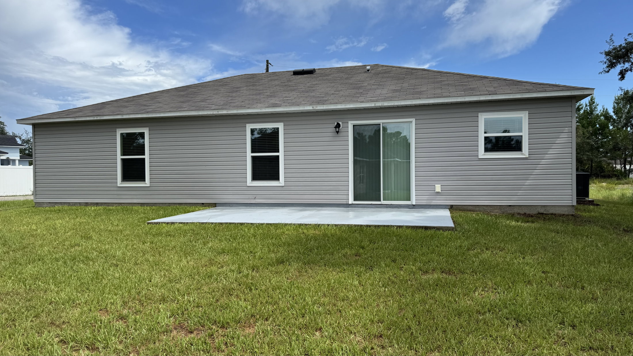Single-story house with a gray exterior and large sliding glass door, set on a grassy lawn under a partly cloudy sky.
