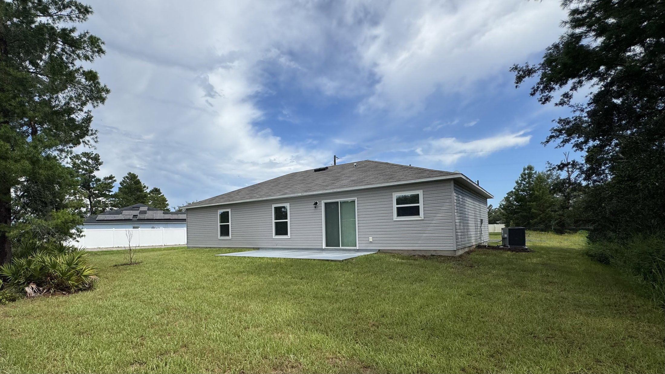 Gray single-story house with a back patio and large grassy area under a partly cloudy sky.