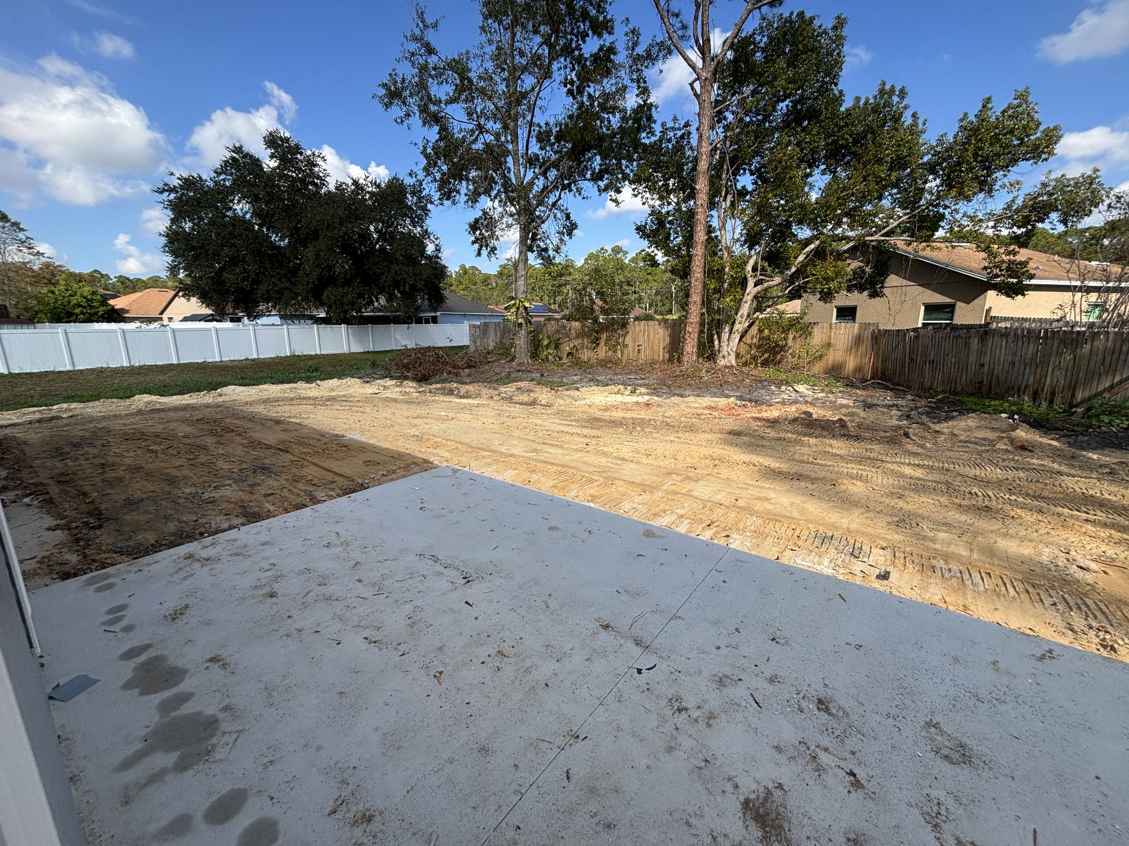 Backyard with a partially paved patio and freshly leveled ground surrounded by trees and a wooden fence, under a clear blue sky.